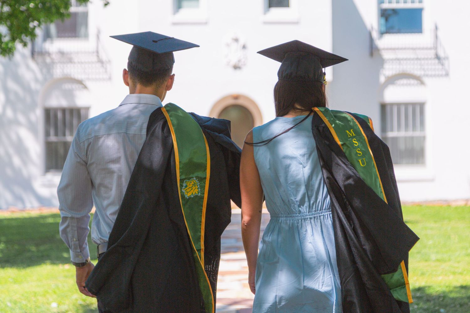 Two graduates in black caps and gowns with green stoles walking together on sunny campus path.
