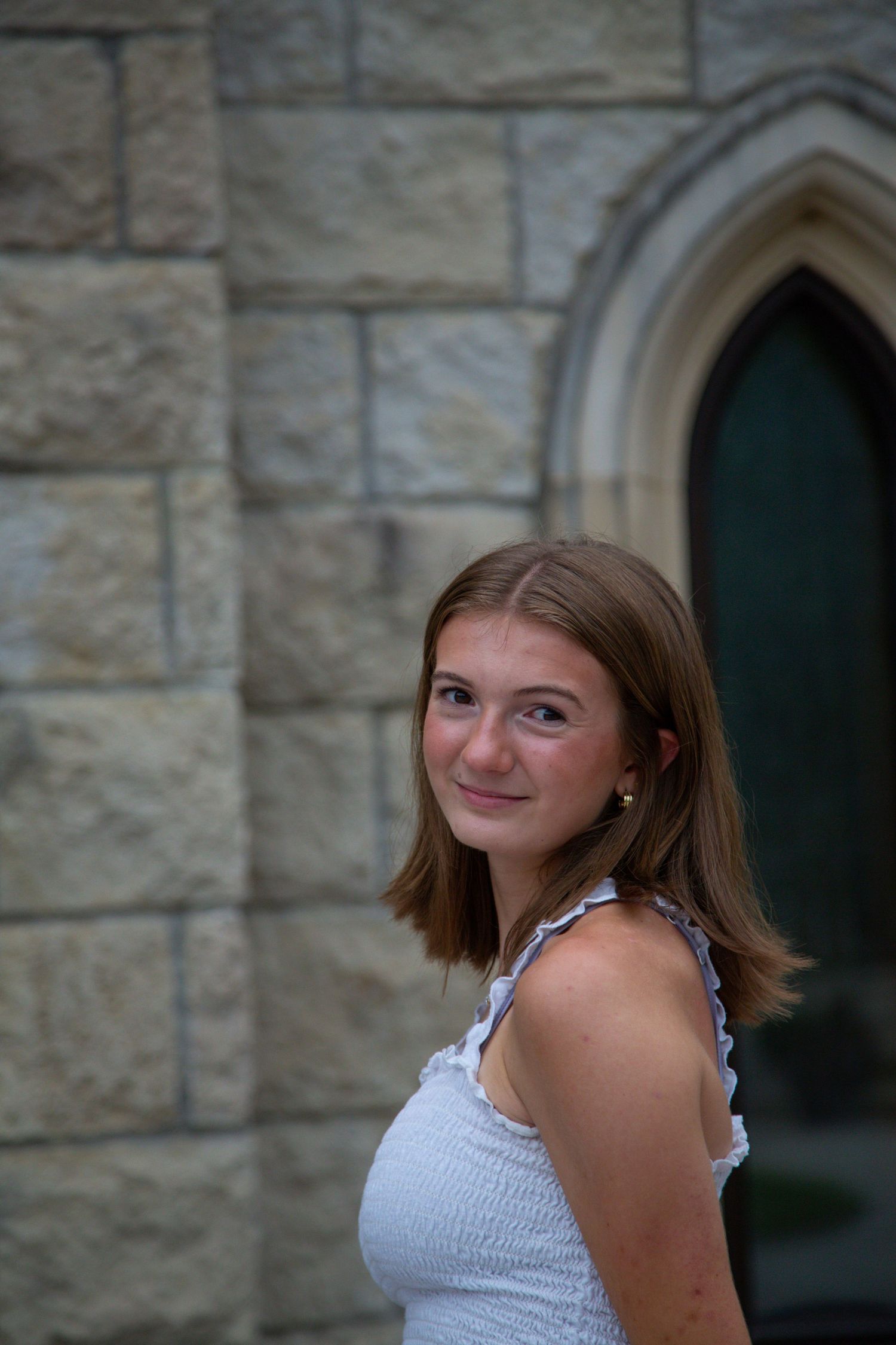 Teen girl with shoulder-length brown hair posing by a gothic stone church wall.