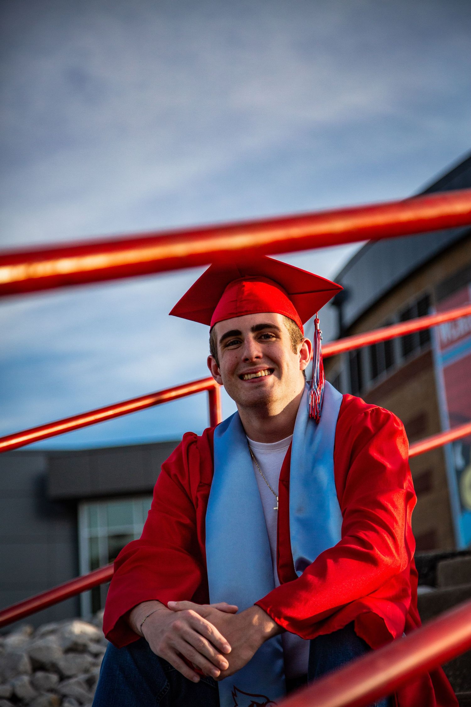 Smiling male graduate in red cap and gown sits on bleacher steps against blue sky.
