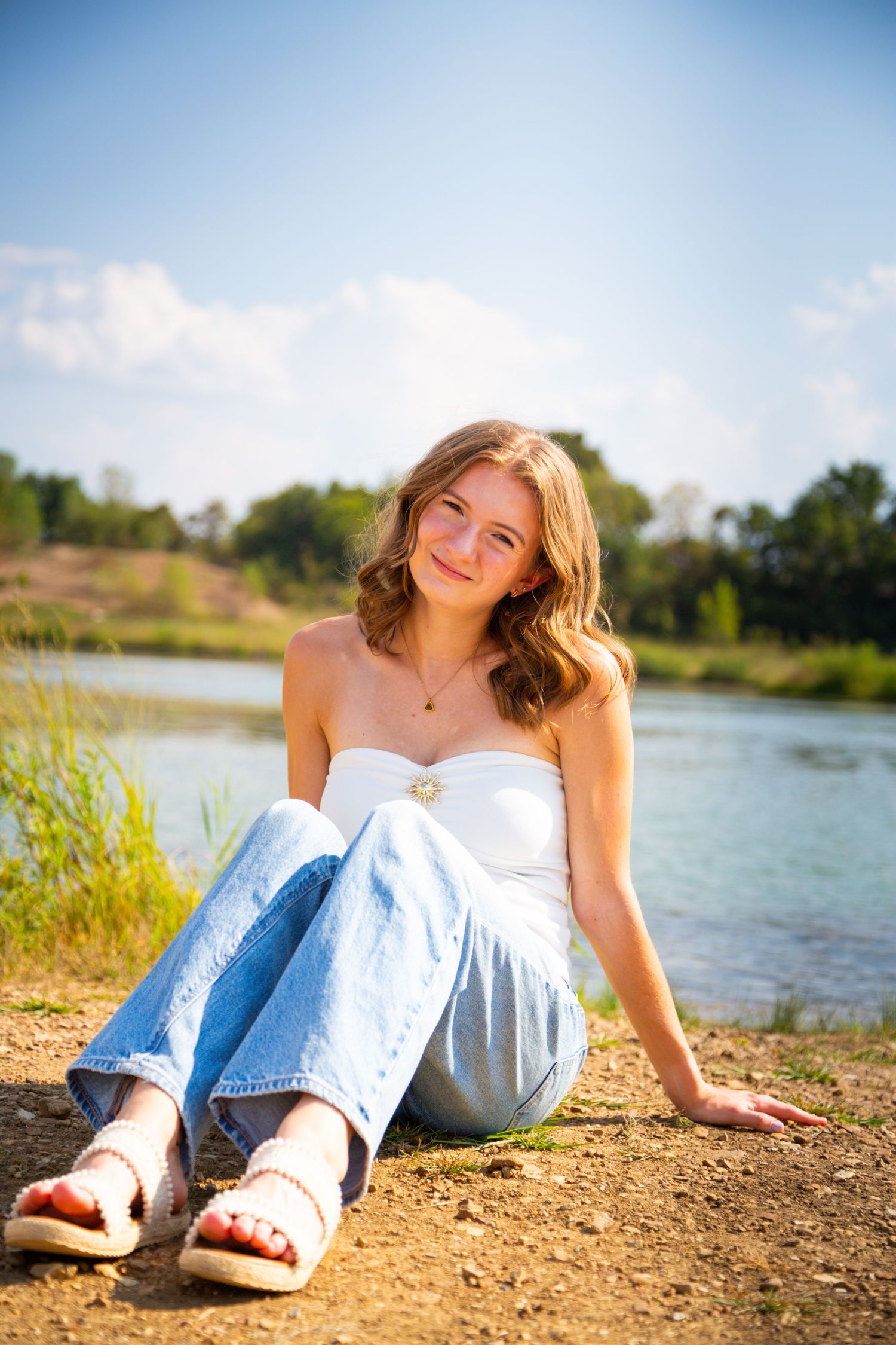 Young woman in white tube top and jeans sitting by a river on a sunny day, smiling outdoors.