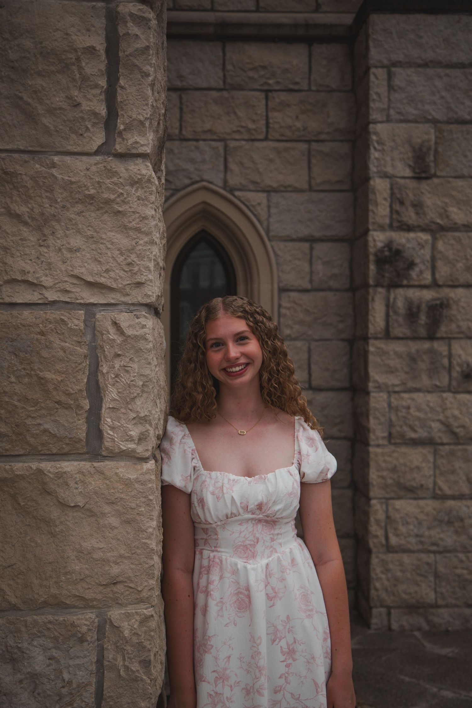 Curly-haired girl in floral dress smiling against a stone gothic church wall.