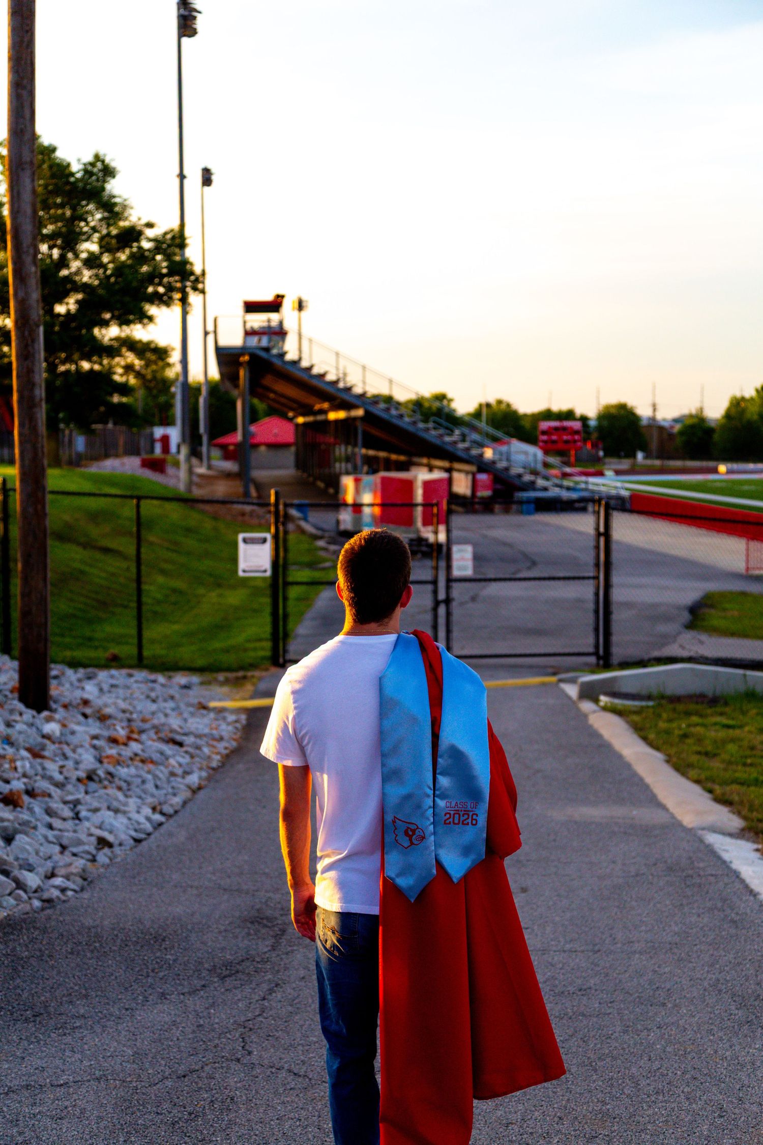 Graduate in white shirt carries red gown and blue stole walking toward stadium at sunset.