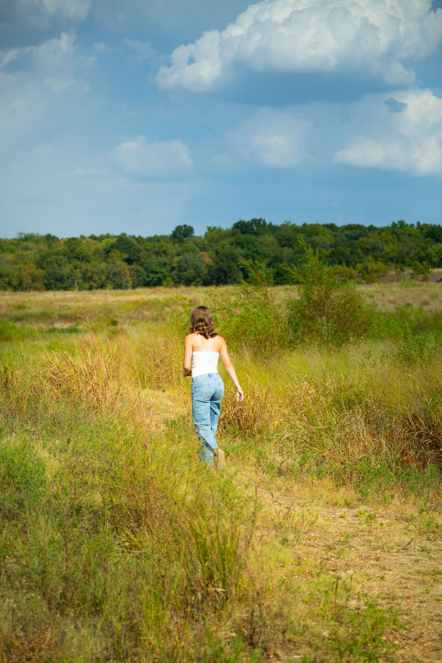 Young woman in white top and jeans walking through a green meadow under a blue cloudy sky.