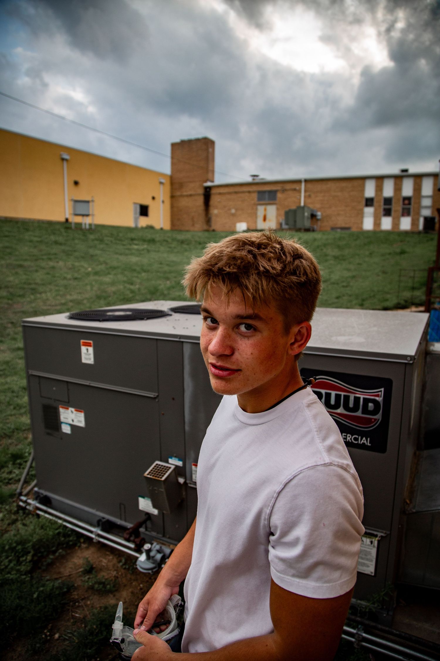Teen boy in white t-shirt stands near commercial HVAC units under dramatic cloudy sky.