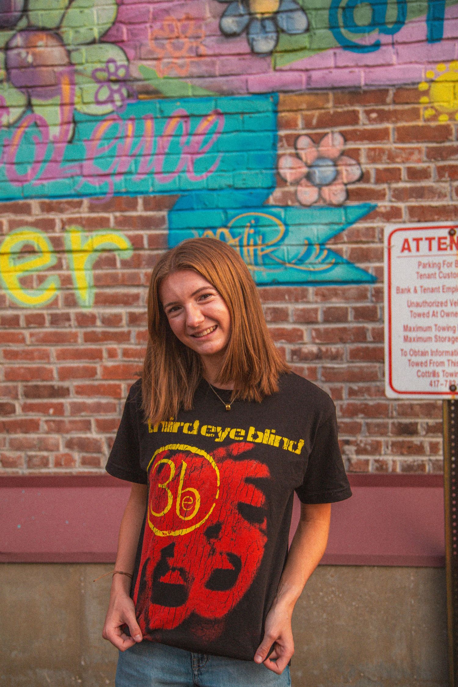 Smiling brunette teen in Third Eye Blind black tee poses before colorful brick wall graffiti mural.