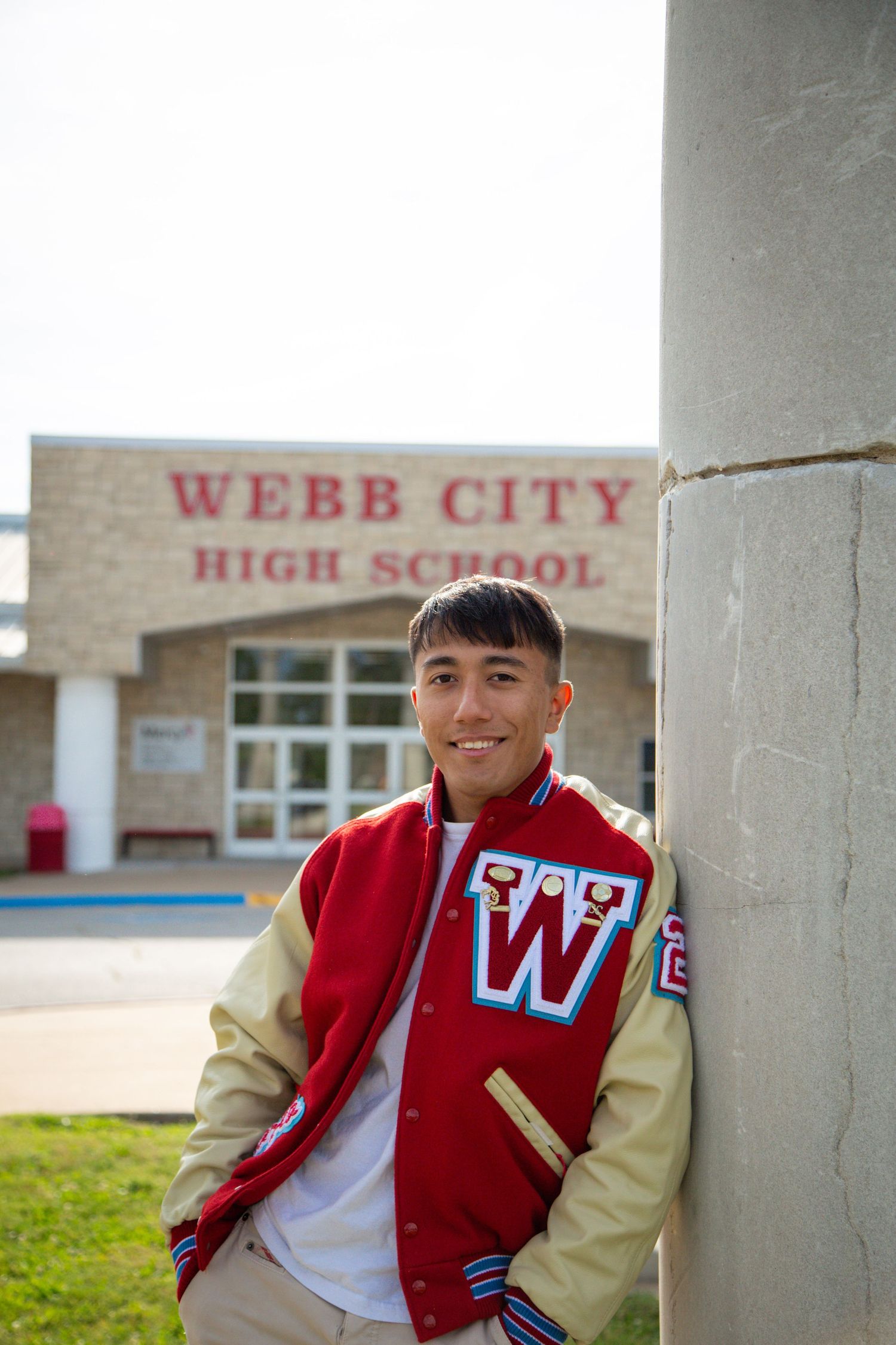 A smiling young man in a Webb City High School varsity jacket leans against a column outside school.