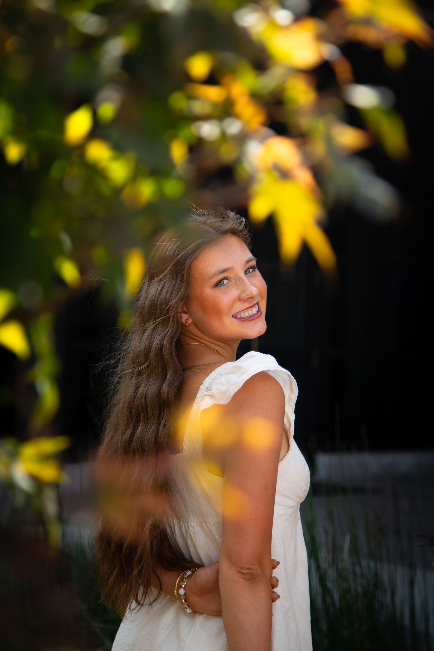 Young woman in white dress smiling outdoors among golden autumn leaves in warm sunlight.