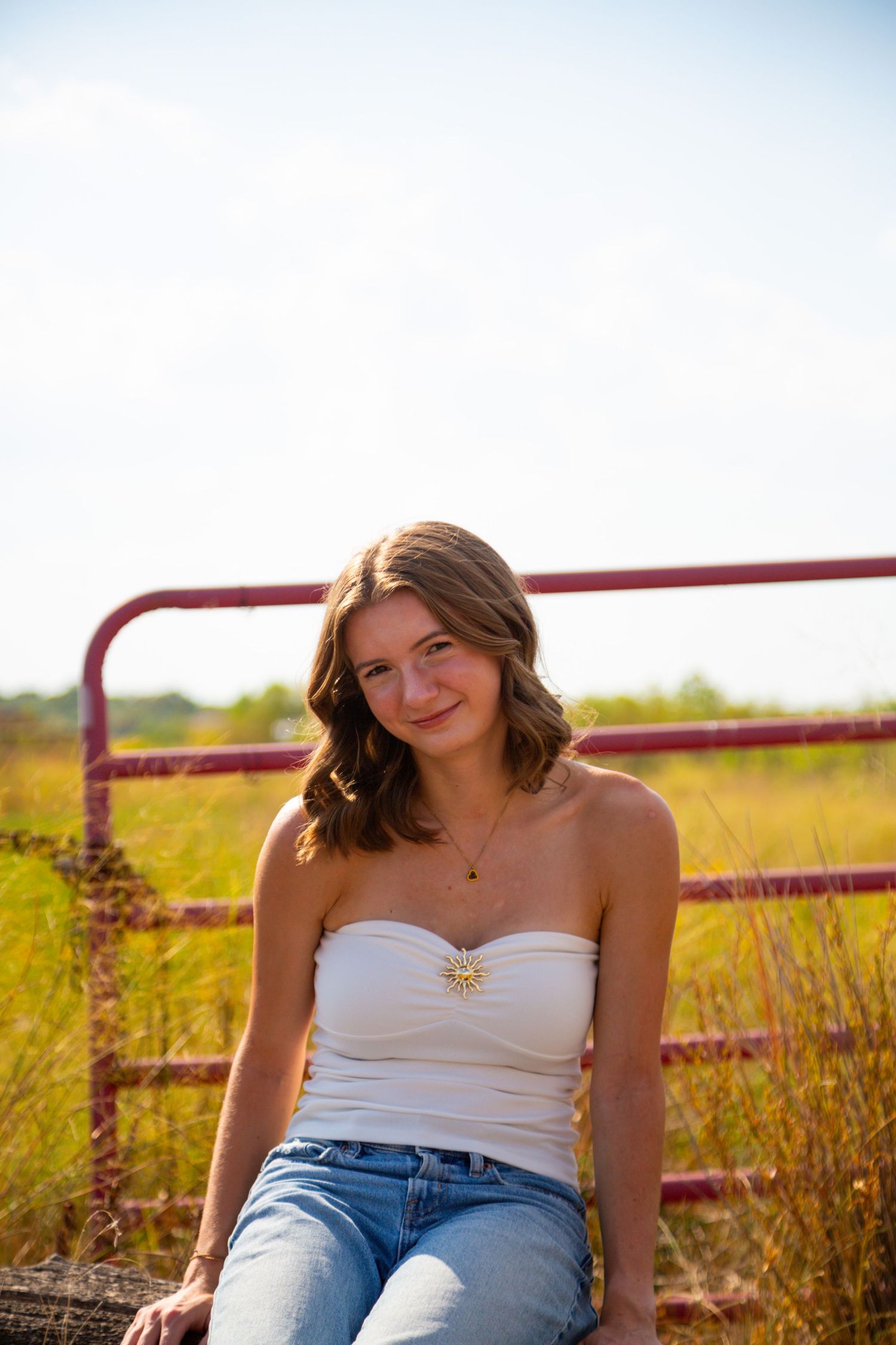Young woman in white tube top and jeans sitting by a rustic red gate in a sunny field.