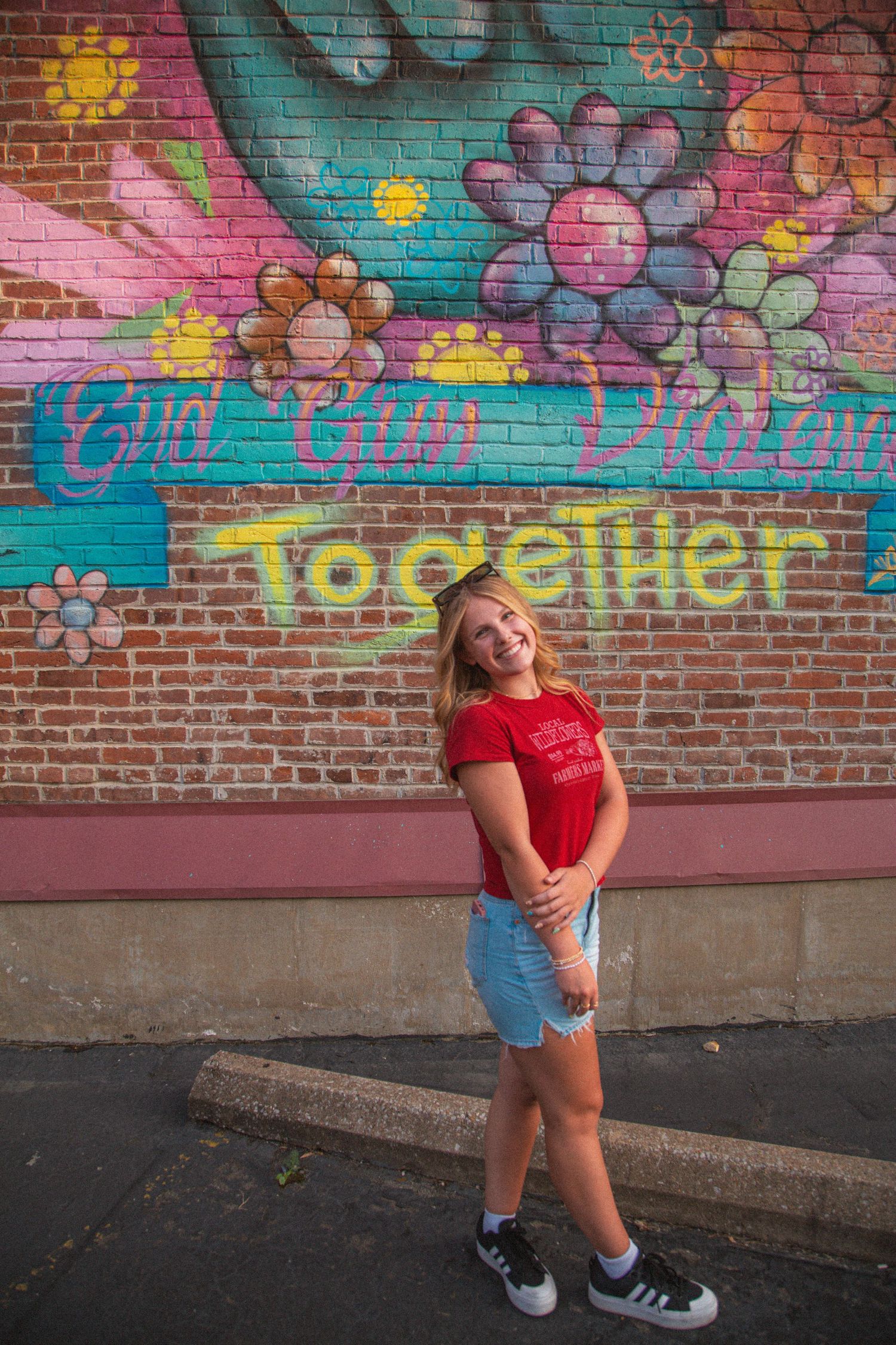 Smiling blonde girl in red tee and denim shorts poses before colorful Together graffiti brick wall.