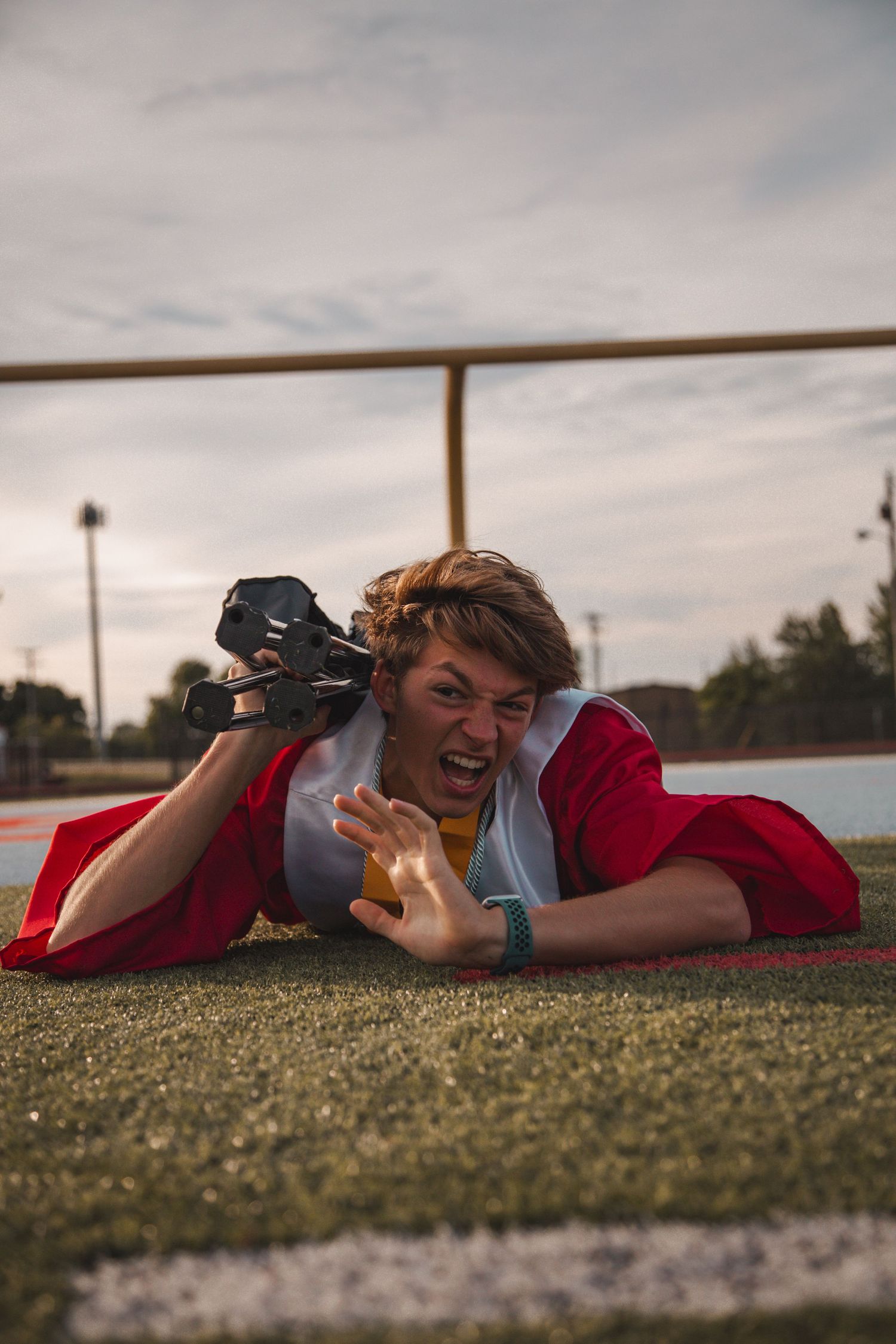 Graduate in red gown lies on turf field holding camera with dramatic expression.