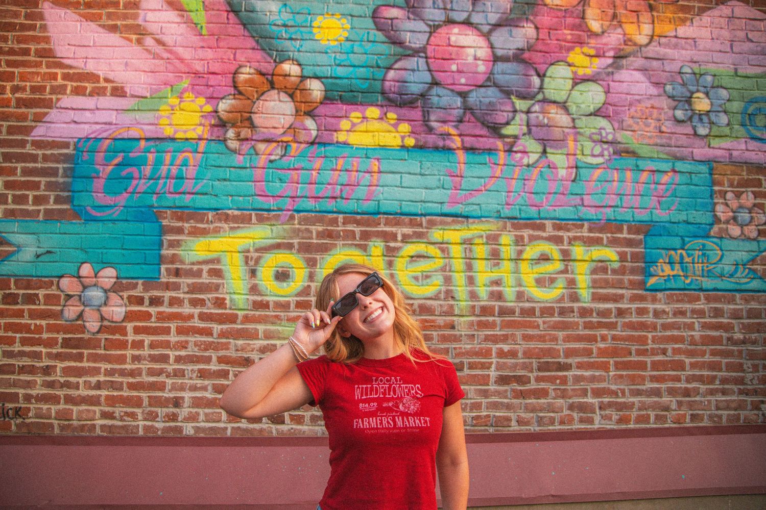 Smiling blonde woman in red Wildflowers Farmers Market tee poses before colorful Together mural.