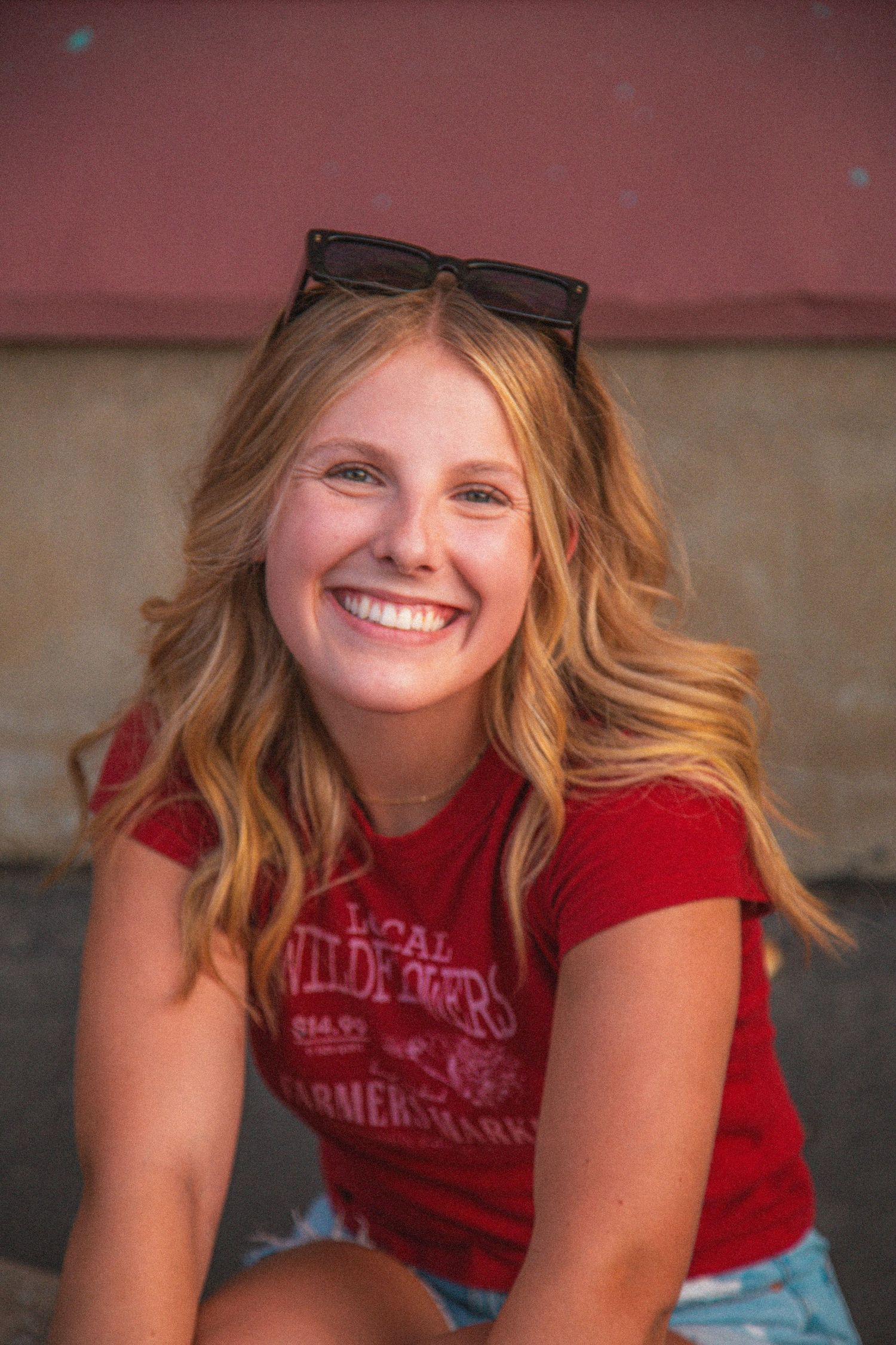 Smiling blonde teen girl in red t-shirt with sunglasses on head outdoors.