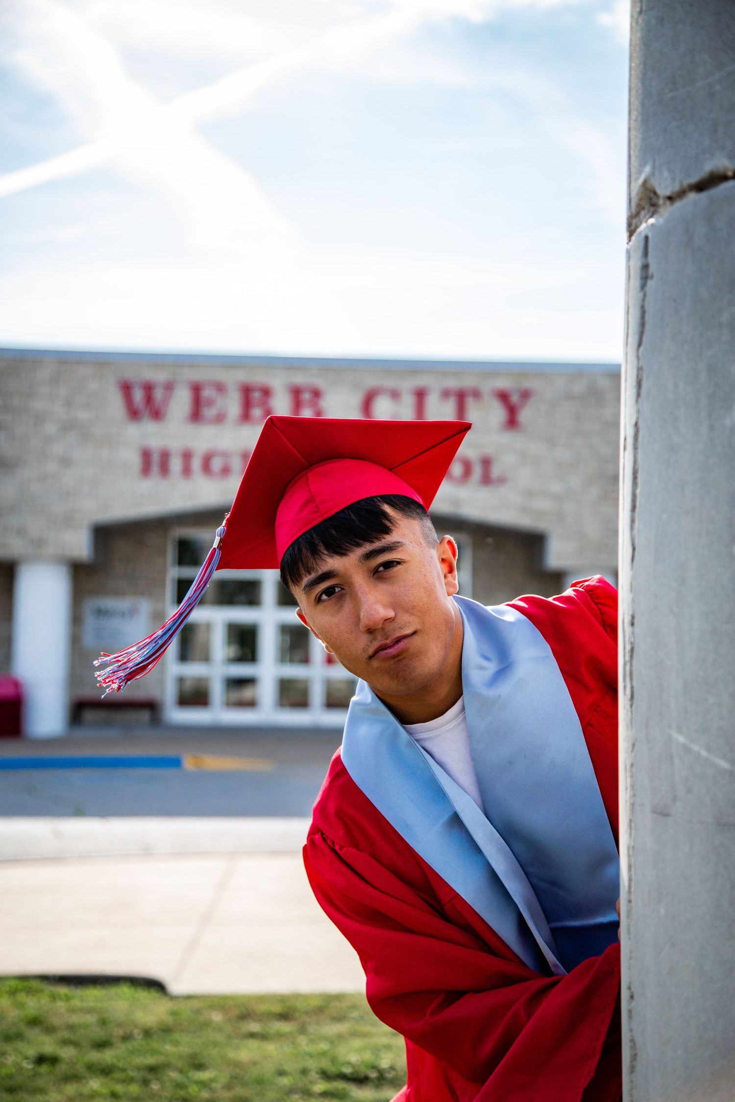 Graduate in red cap and gown leaning against pole at Webb City High School.