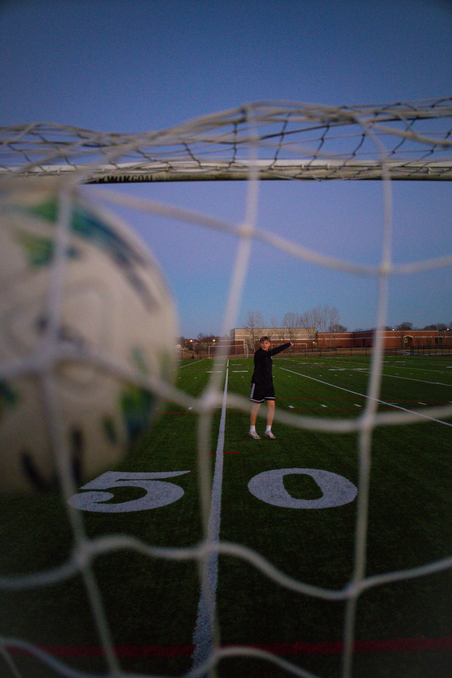 Soccer ball in foreground of net with player standing on field at the 50-yard line at dusk.