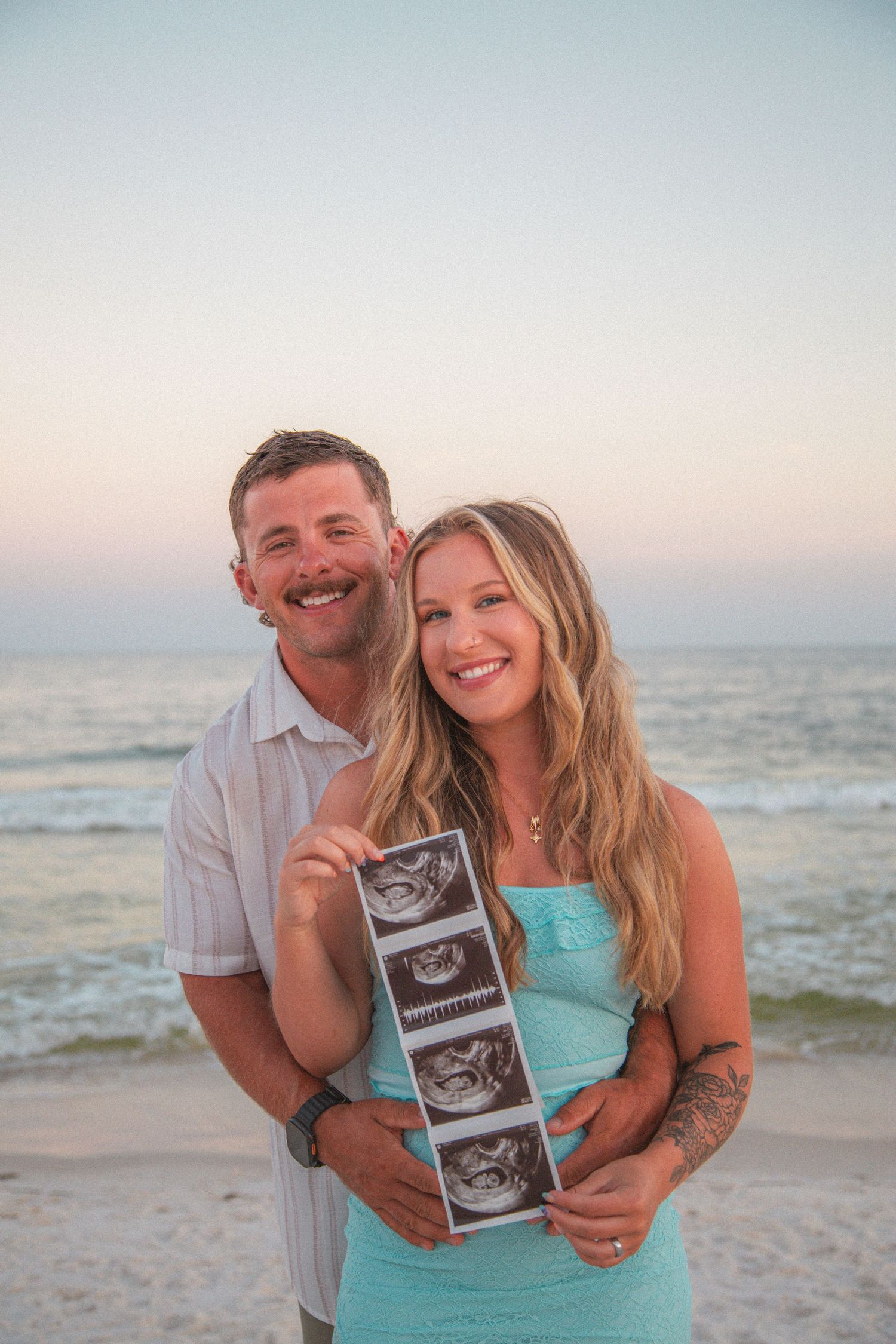 Smiling couple displaying ultrasound strip photos at beach during golden hour pregnancy reveal.