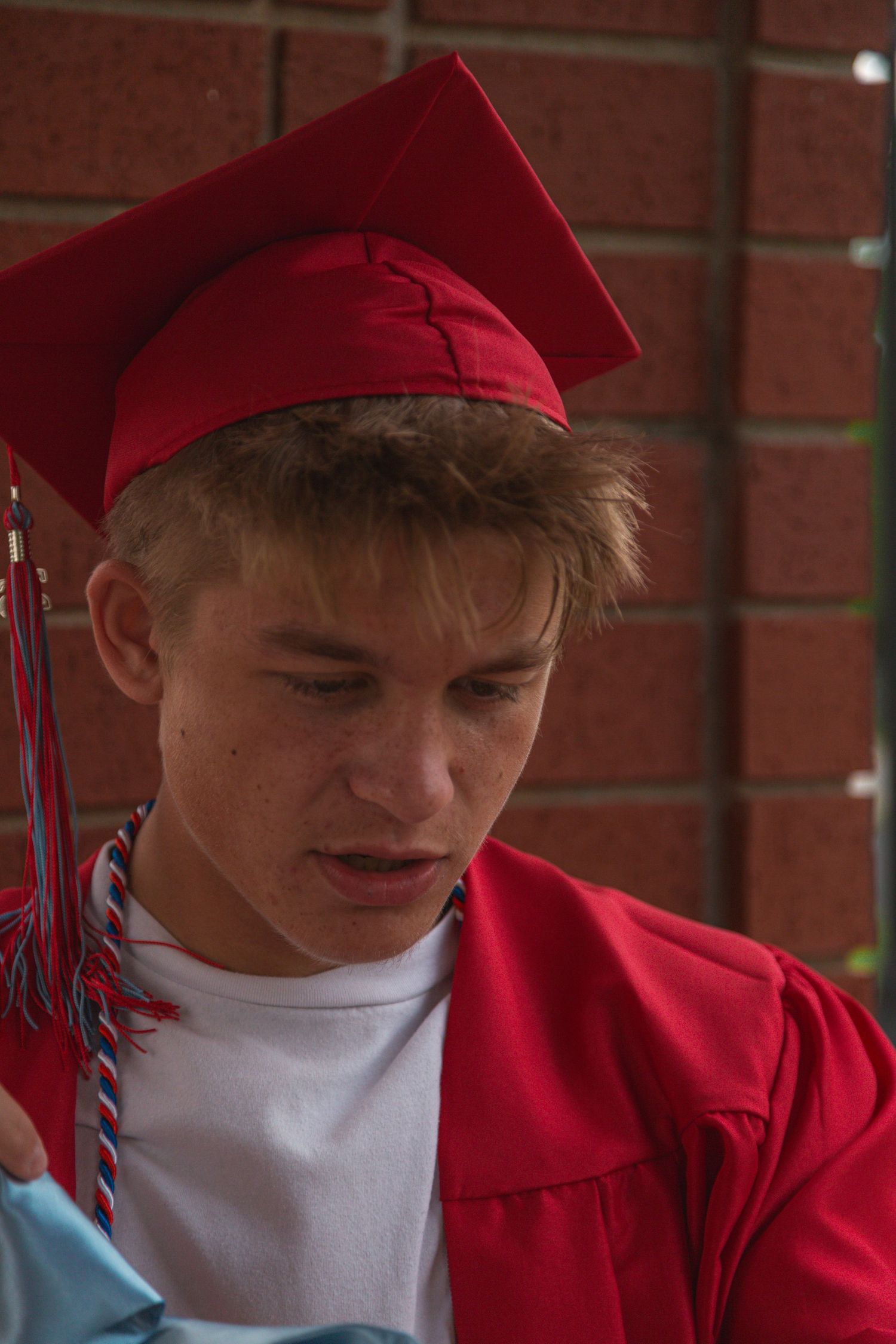 Young man in red graduation cap and gown looking down near brick wall.