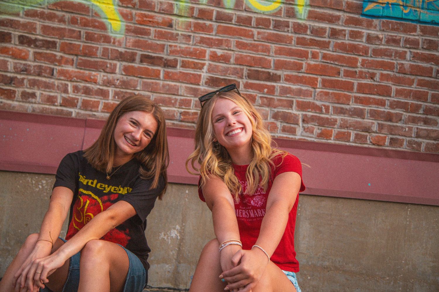 Two smiling teenage girls sitting together against a brick wall with colorful graffiti in warm sunlight.