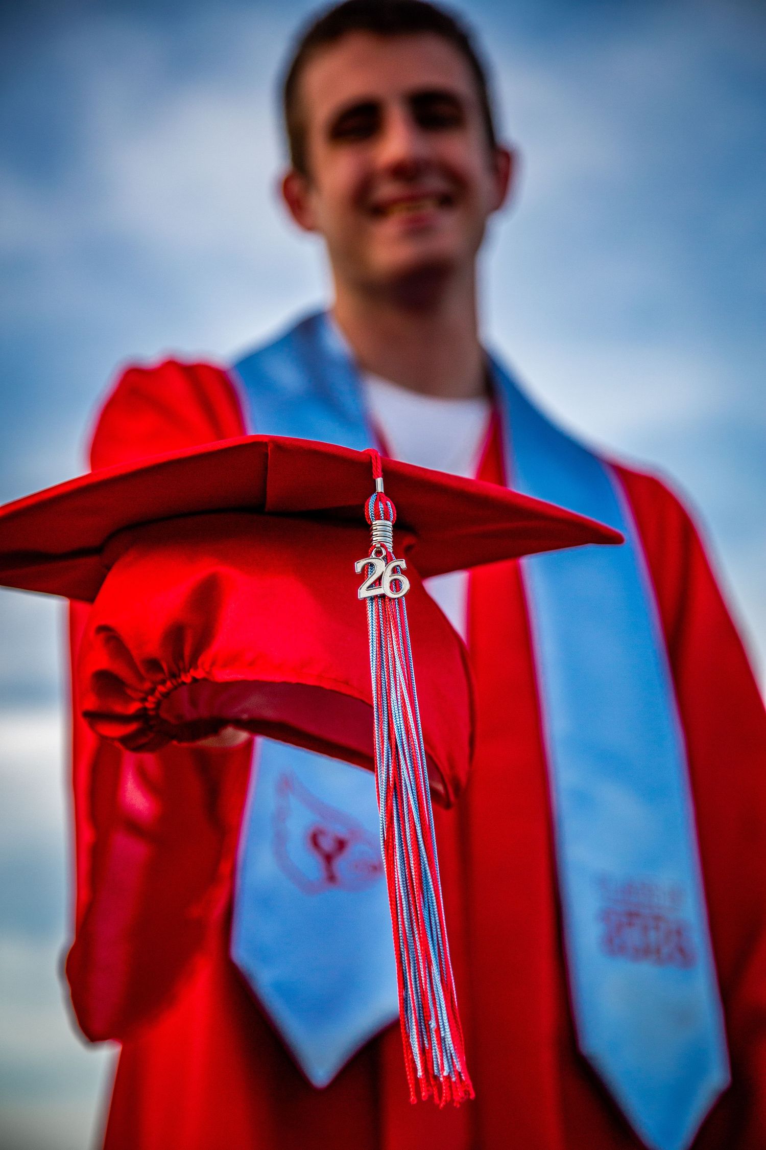 A smiling graduate in red gown and light blue stole extends his red mortarboard cap with 26 tassel.