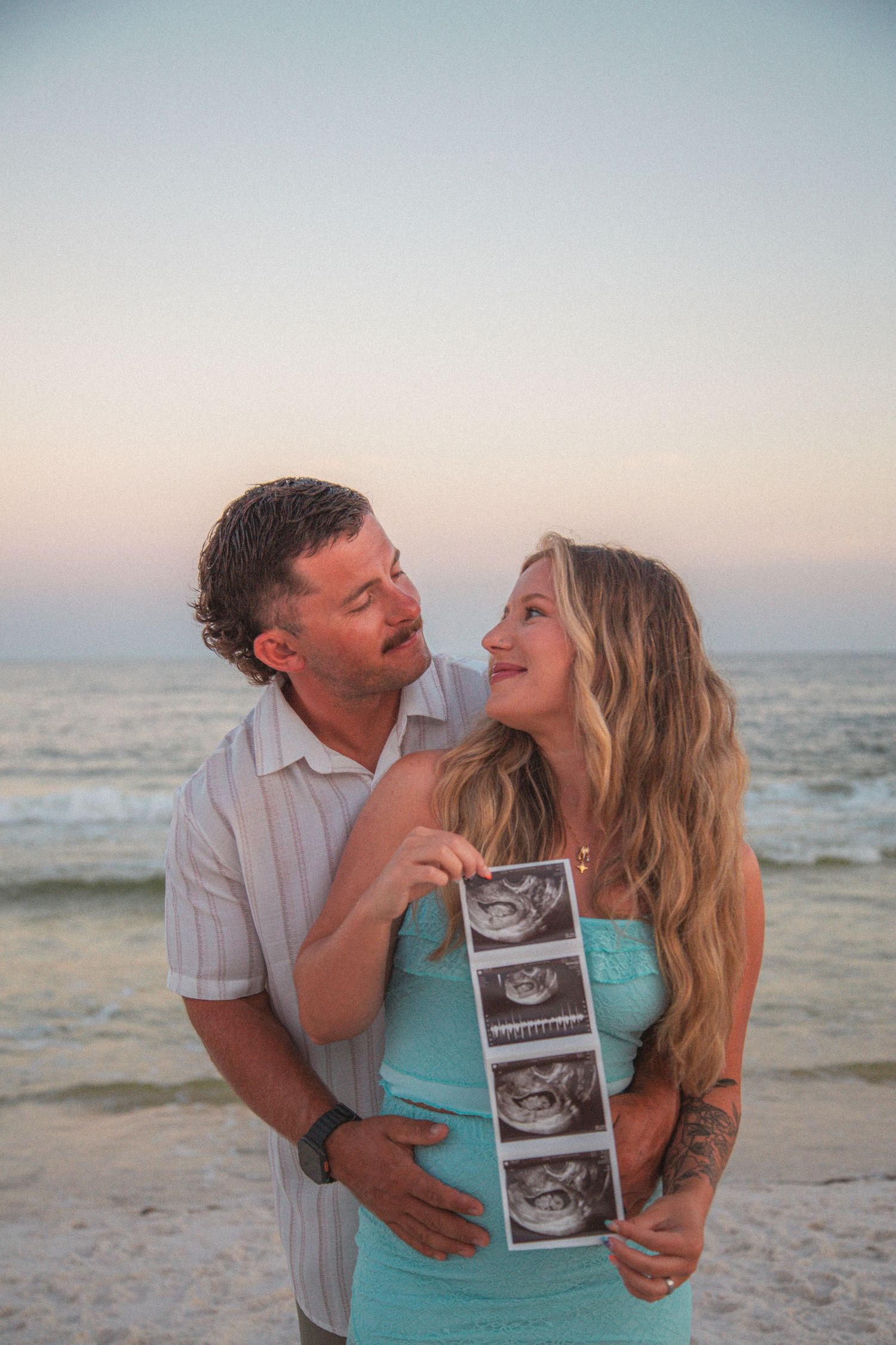 Happy couple holding ultrasound photos at beach sunset, sharing pregnancy announcement moment.