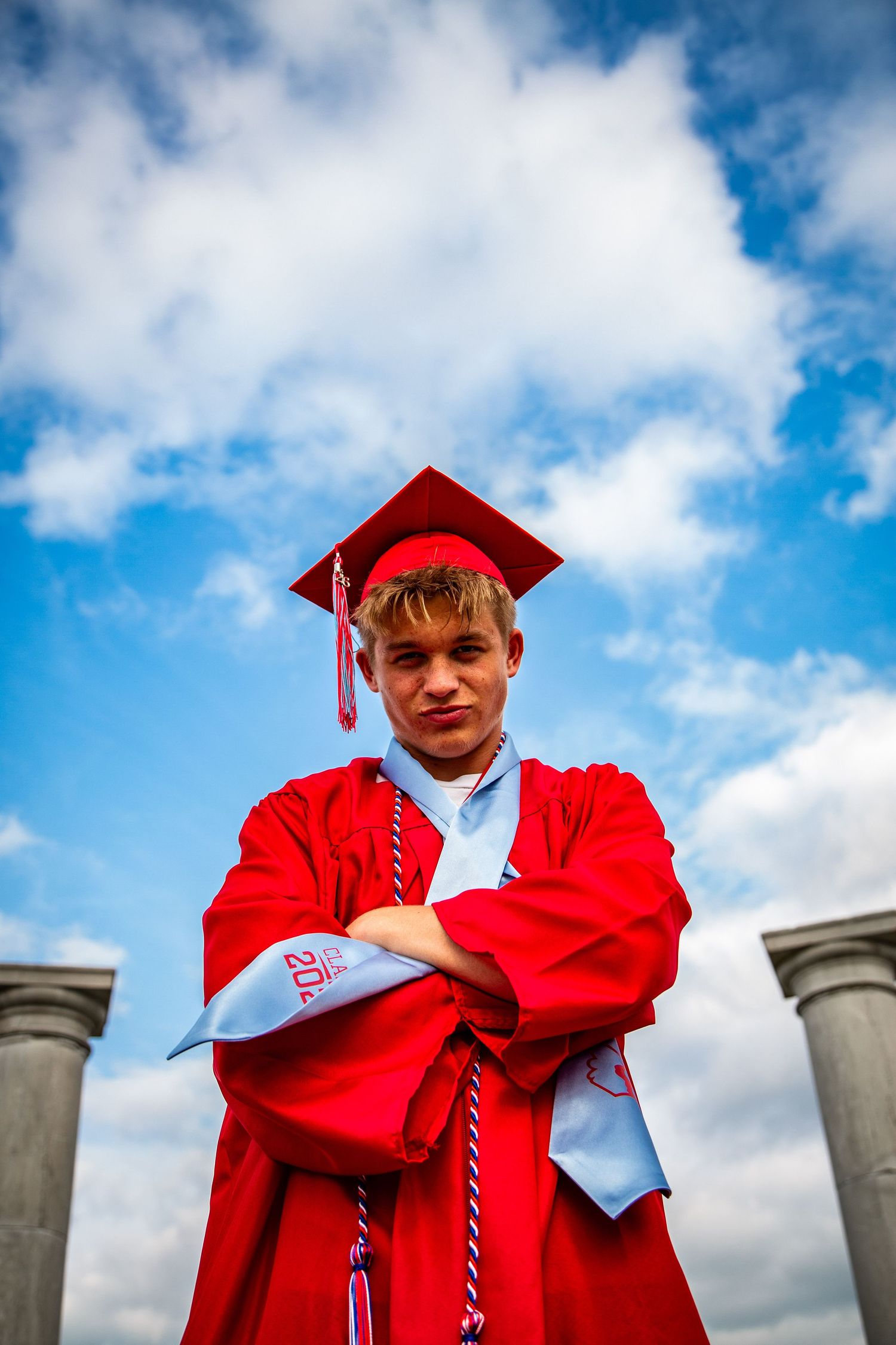 Graduate in red cap and gown posing confidently with arms crossed under blue sky.
