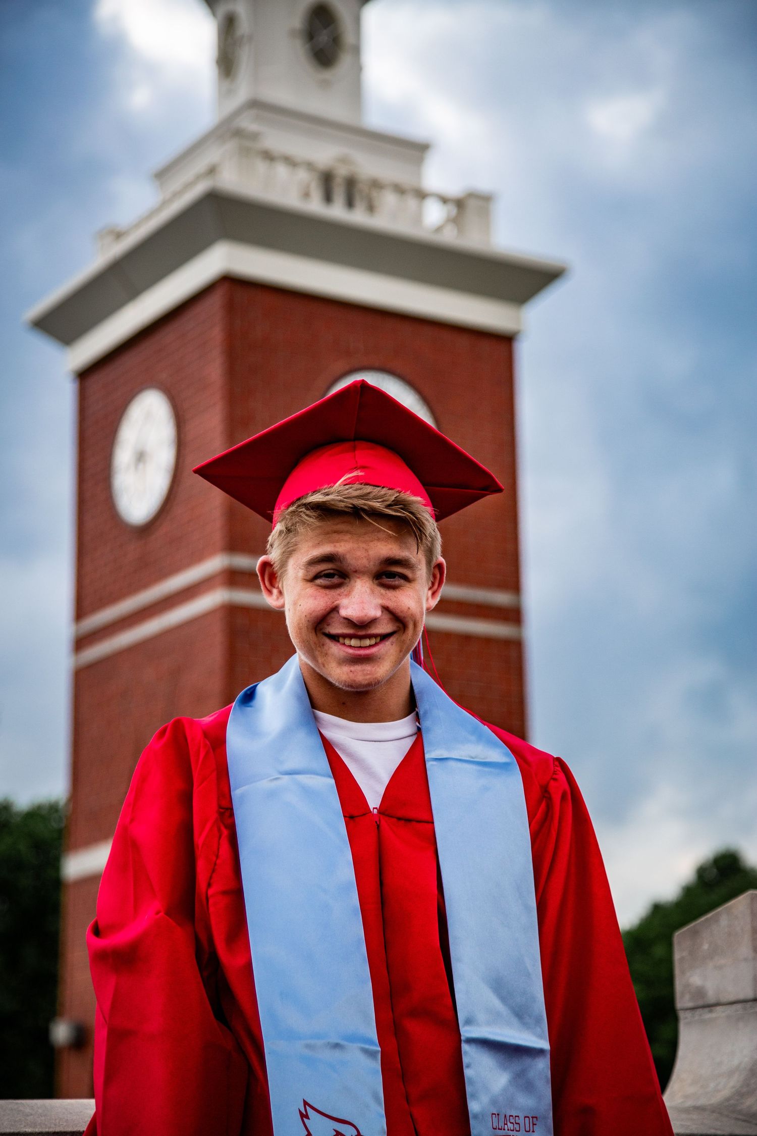 A smiling young male graduate in red cap and gown poses in front of a brick clock tower.
