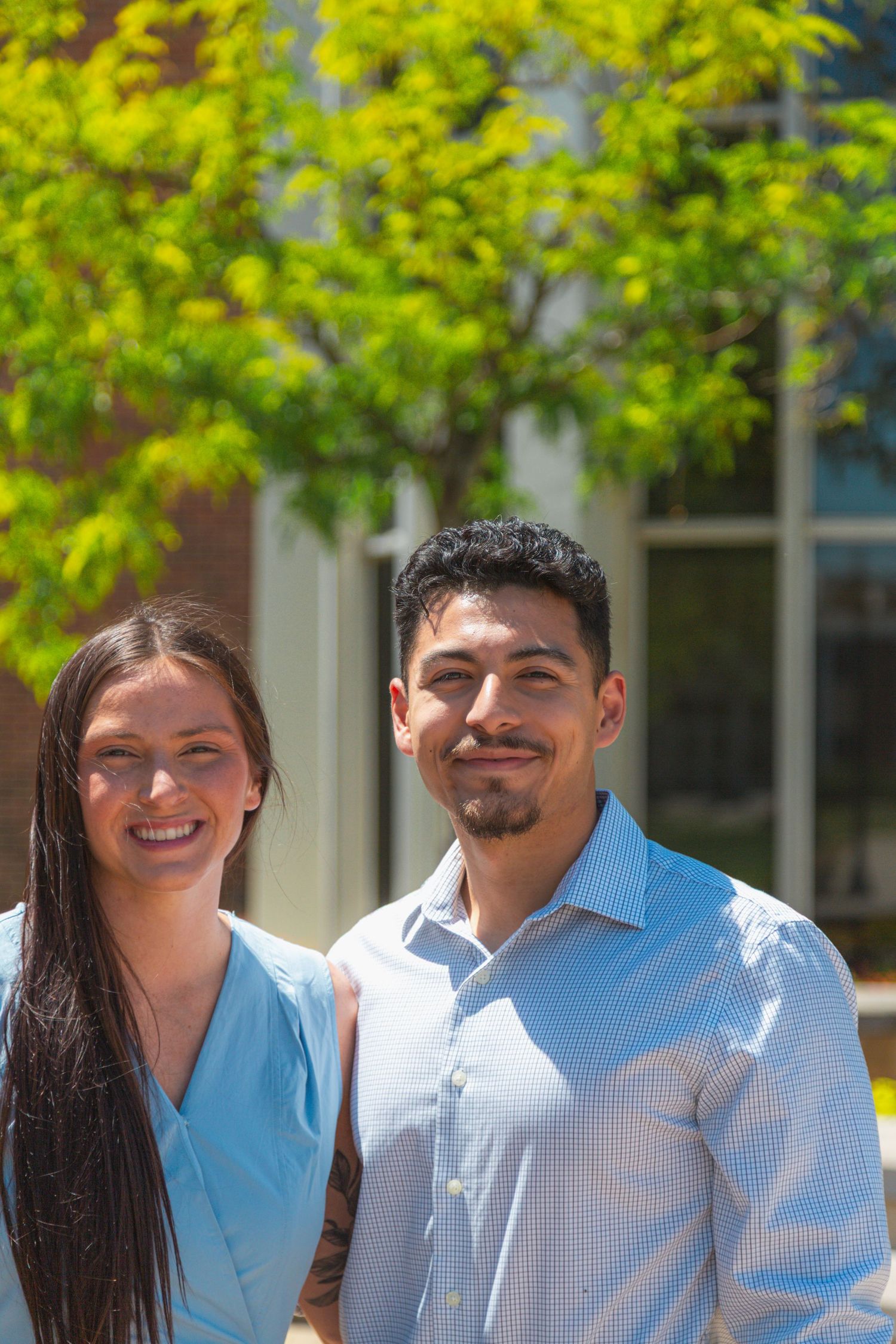 Smiling young couple standing together outdoors under green trees on sunny day.