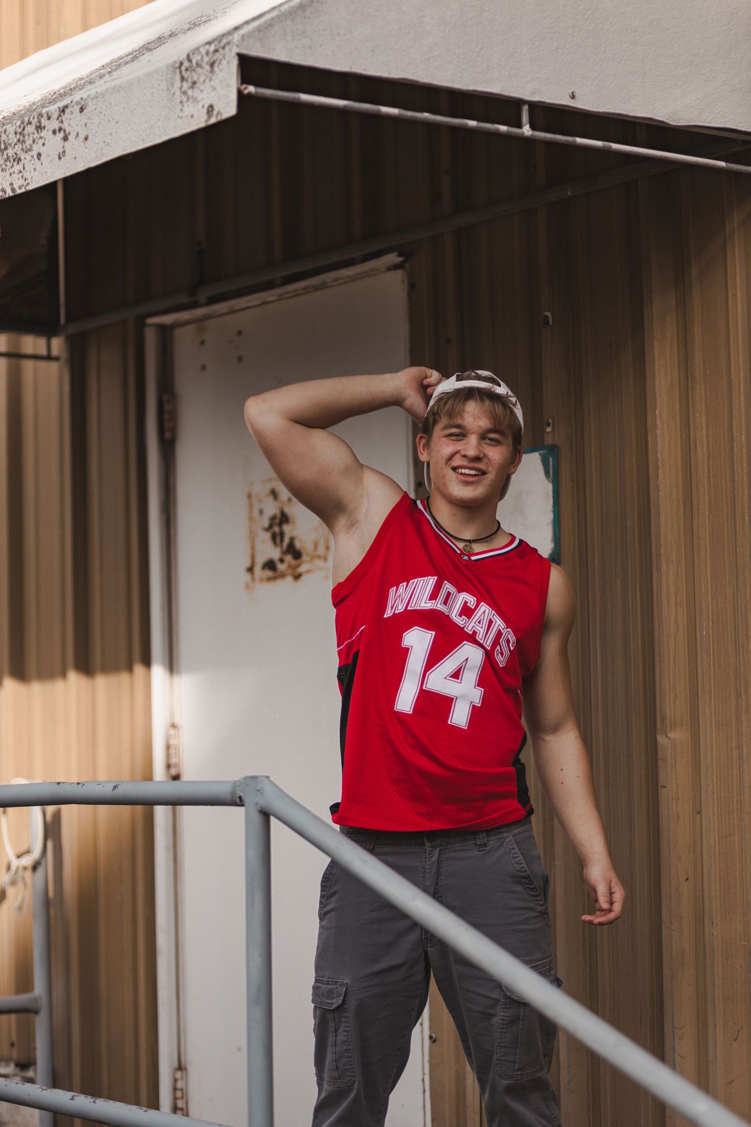 Smiling teen boy in Wildcats number 14 jersey leans on railing of old building.