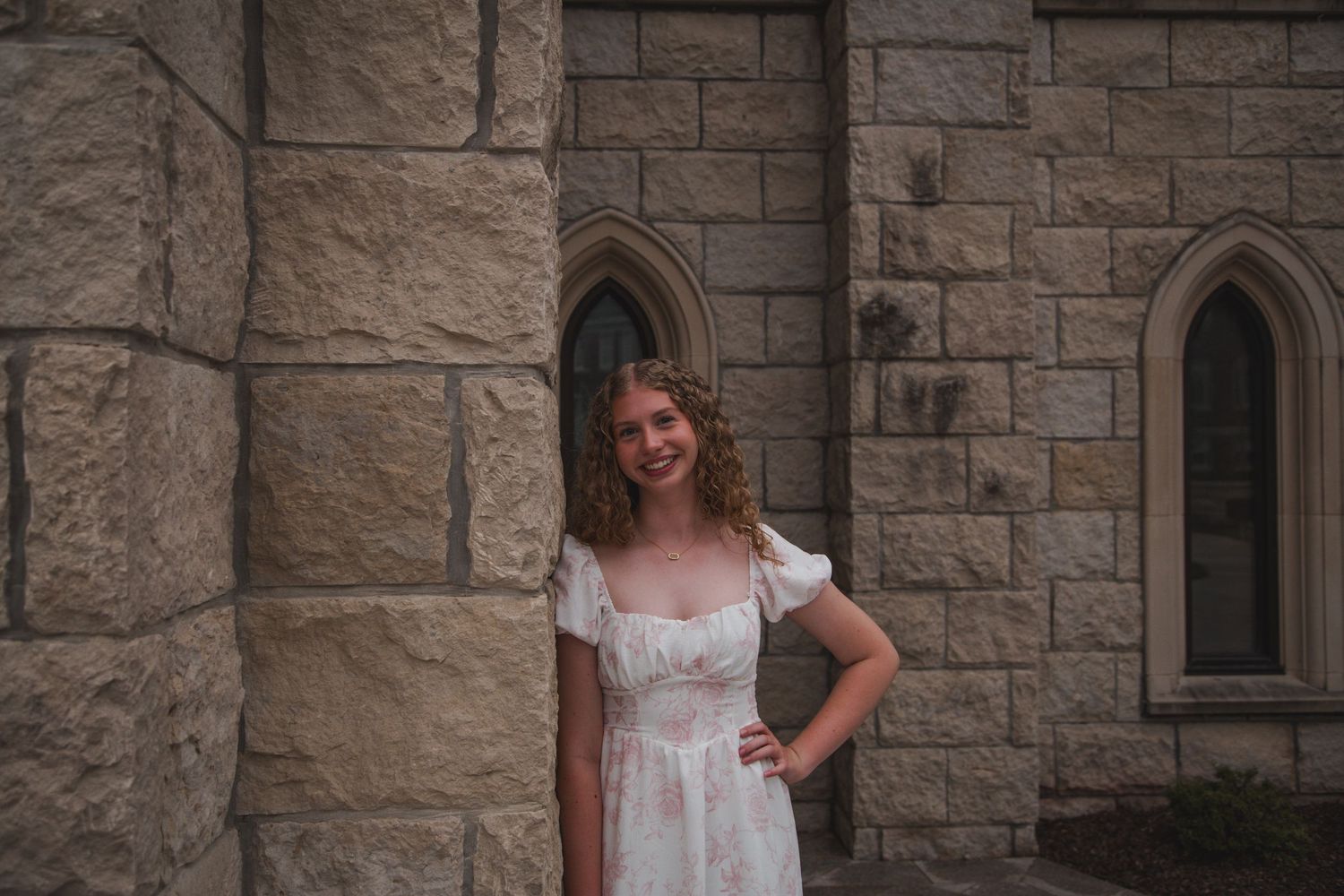 Smiling young woman in floral white dress standing in gothic stone church doorway.