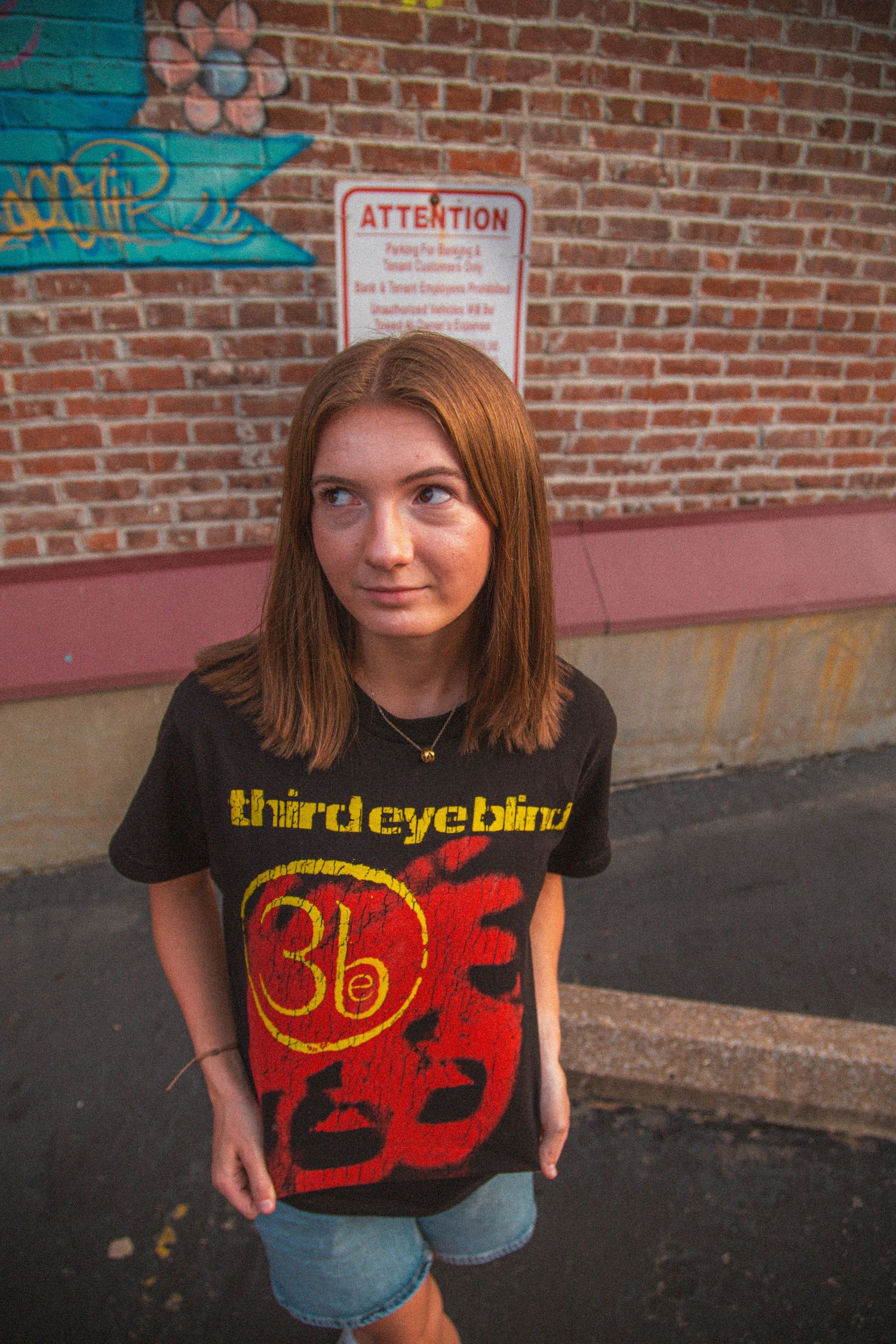 Teen girl in a Third Eye Blind t-shirt stands casually in front of a brick wall at golden hour.