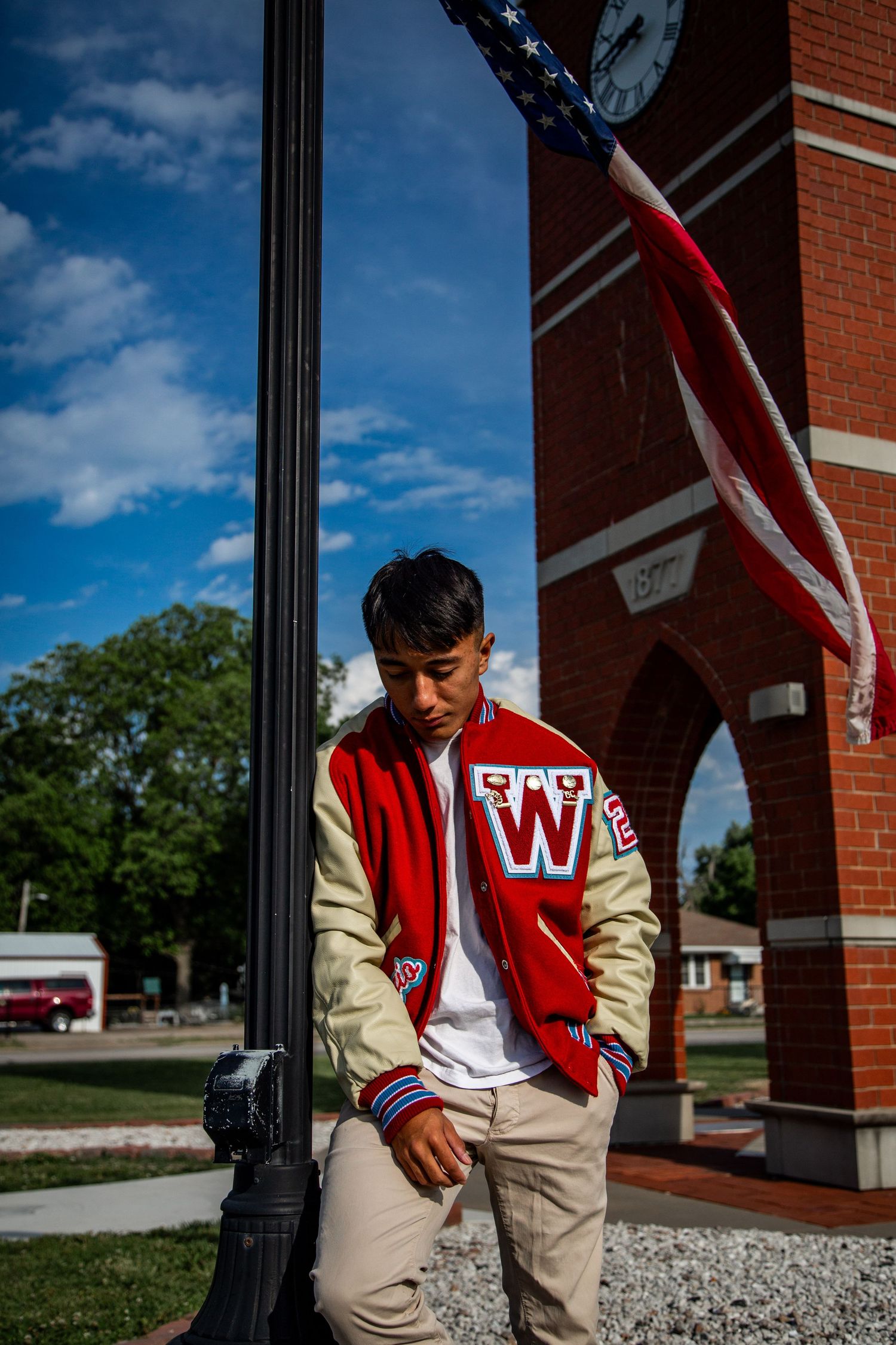 Young man in red varsity jacket with W letter leaning against lamppost near brick clock tower with American flag.