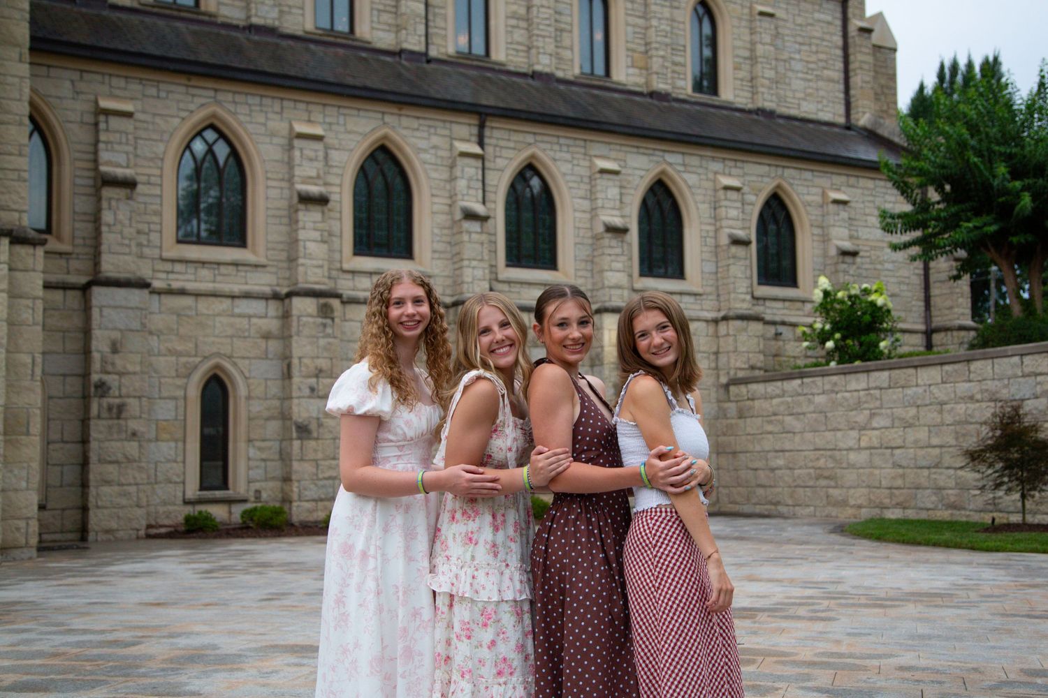 Four young women in elegant dresses smiling together outside stone church.