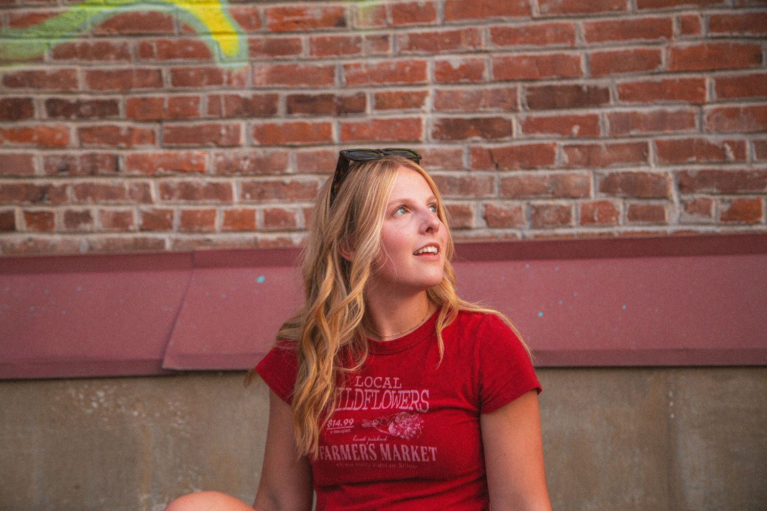 Blonde teenage girl in red t-shirt sits against brick wall looking upward outdoors.