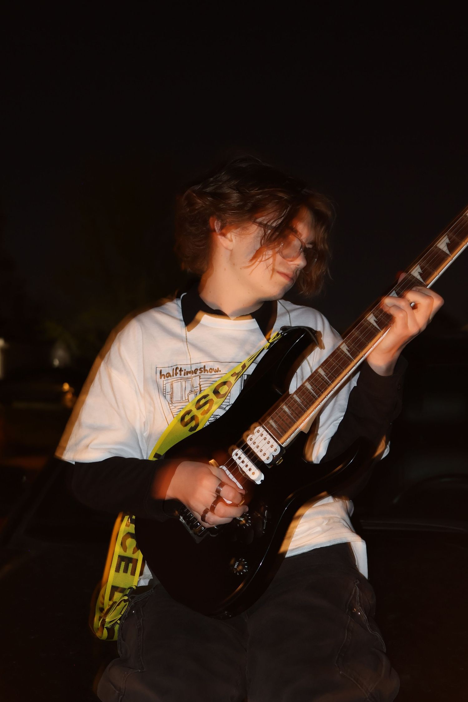 A teenage guitarist plays a black electric guitar outdoors at night wearing a graphic t-shirt.