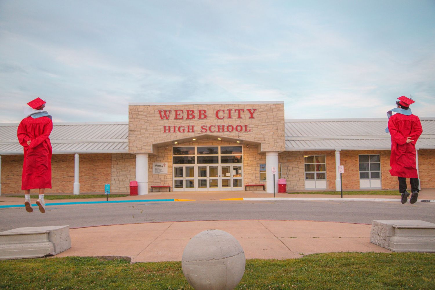 Two graduates in red caps and gowns jumping in front of Webb City High School entrance at dusk.