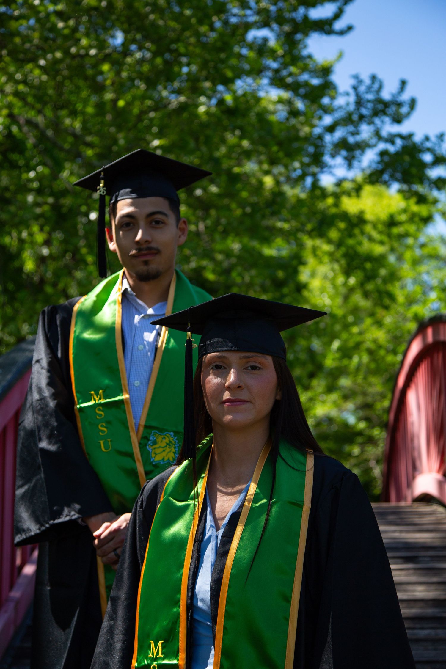Two college graduates in black caps and green MSU stoles posing on red bridge surrounded by trees.