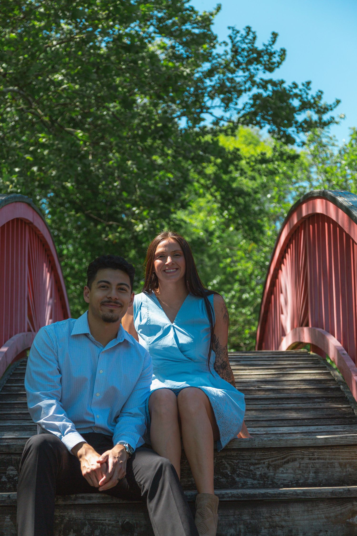 Smiling couple in coordinating blue outfits sitting on wooden steps of red arched bridge outdoors.