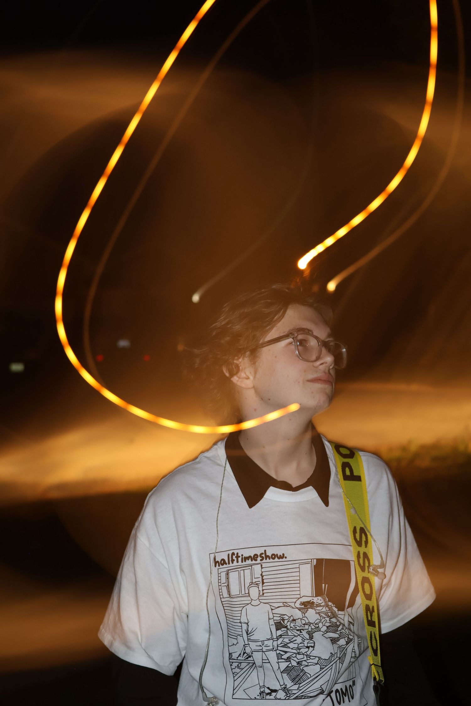 A young man wearing glasses and a graphic tee looks up at glowing light trails in the dark.