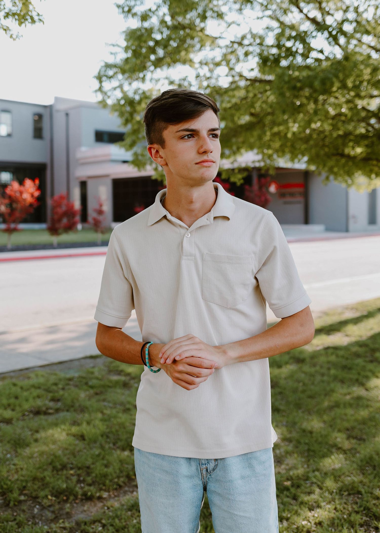 Young man in beige polo shirt stands outdoors near school building among trees.
