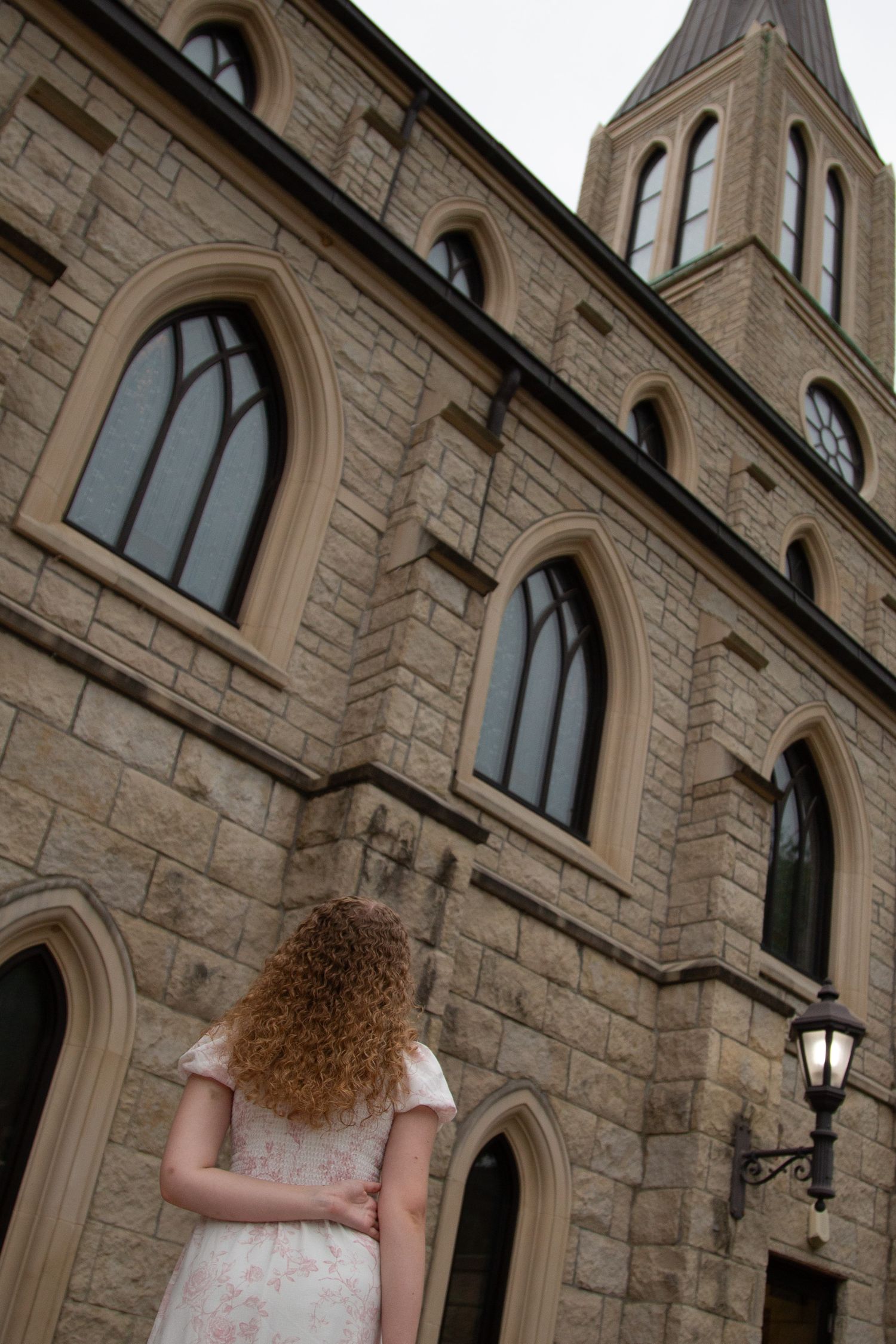 Young woman with curly red hair in floral dress gazing up at stone church.