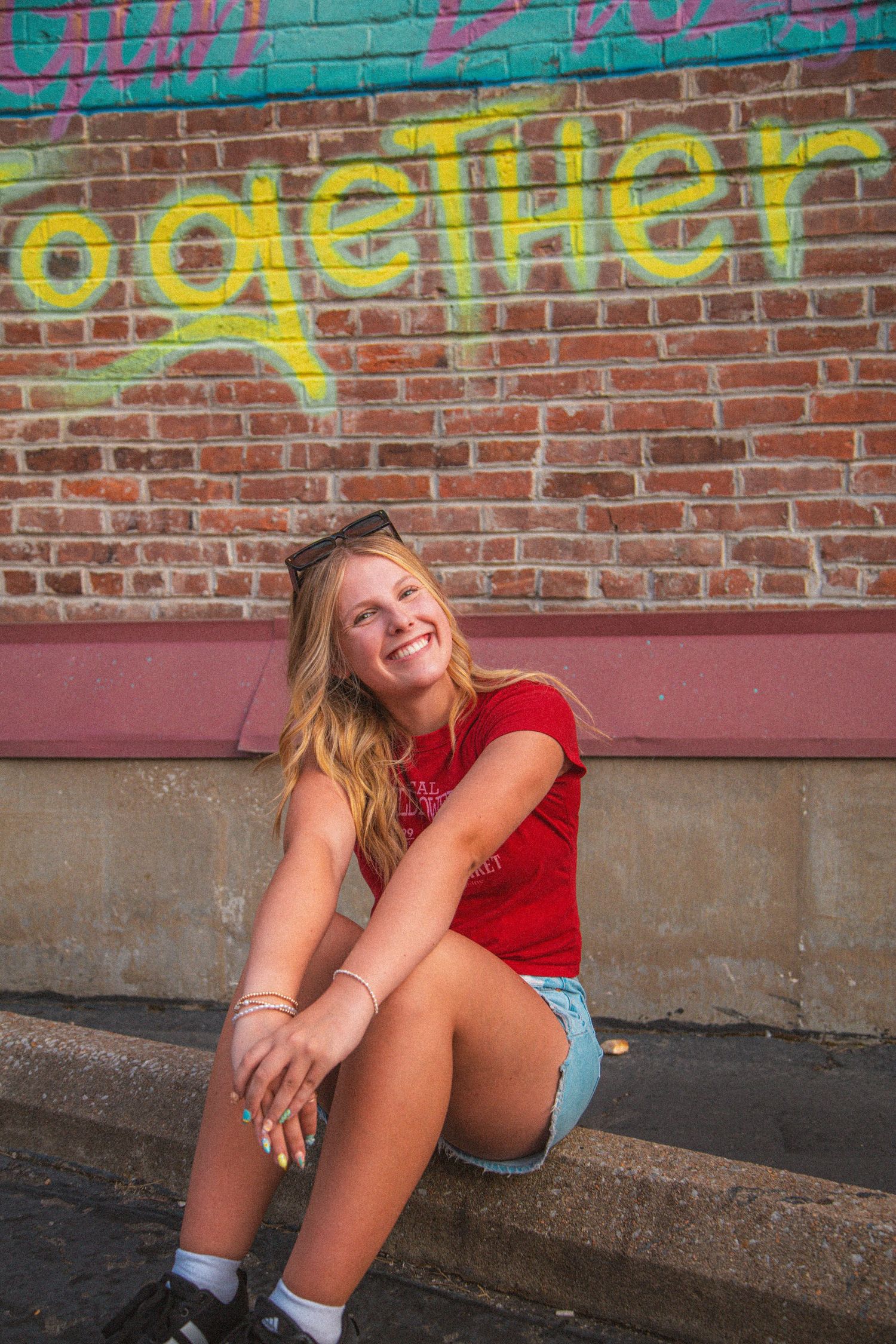Smiling blonde teen in red tee sits against brick wall with yellow Together graffiti mural behind.