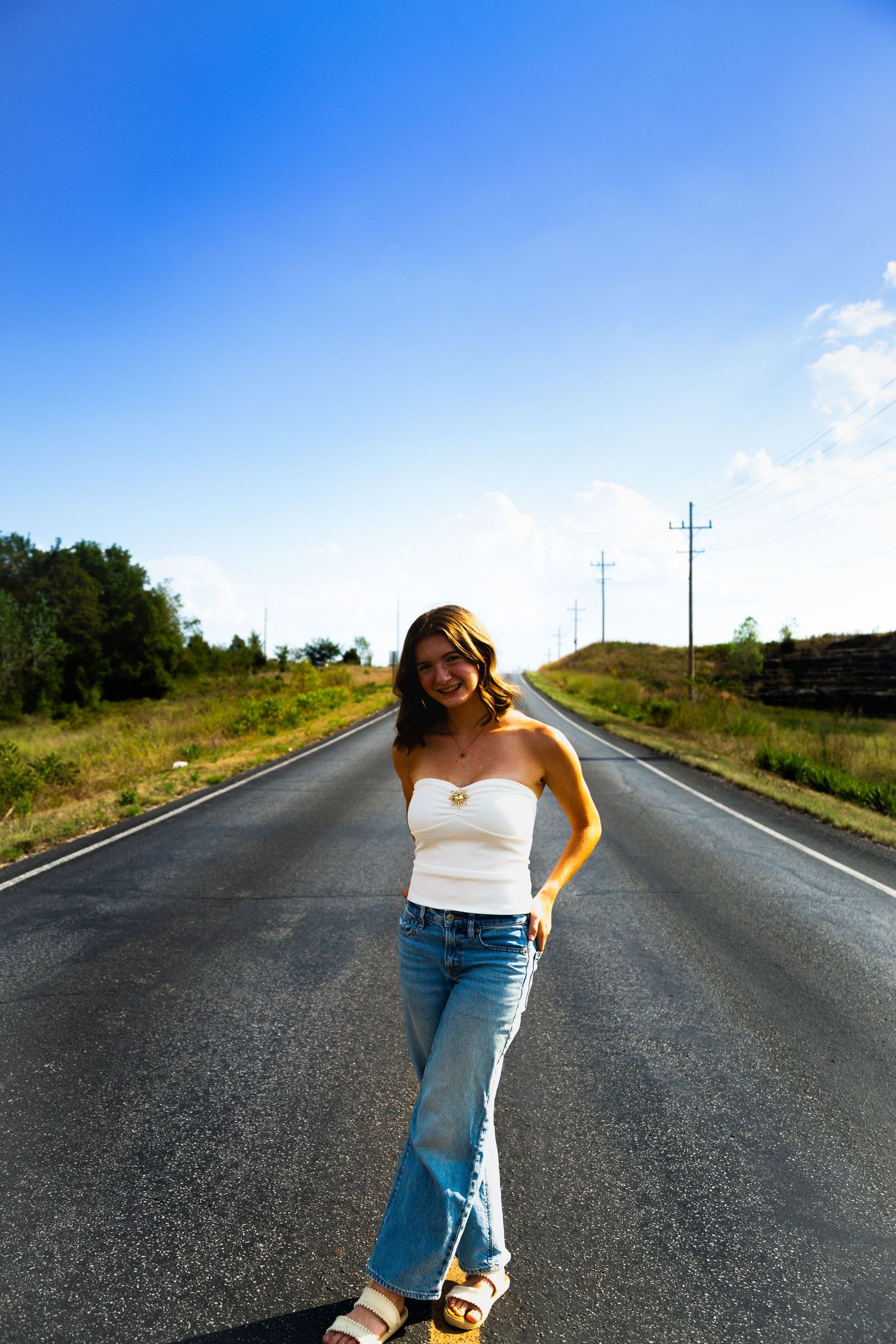 Young woman in white tube top and jeans standing on an empty road under a bright blue sky.