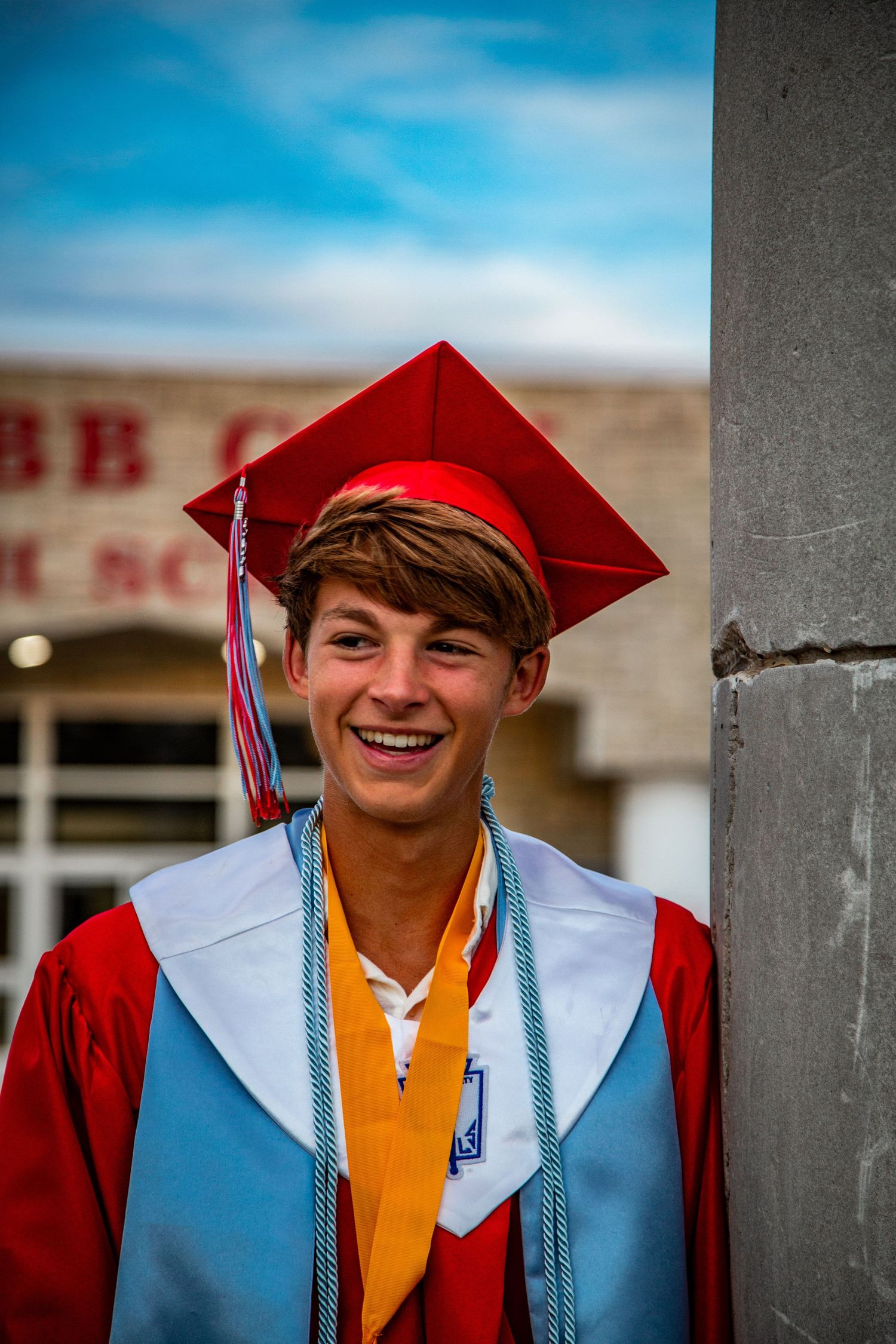Smiling young male graduate in red cap and gown leaning against a pillar outside Webb City High School.