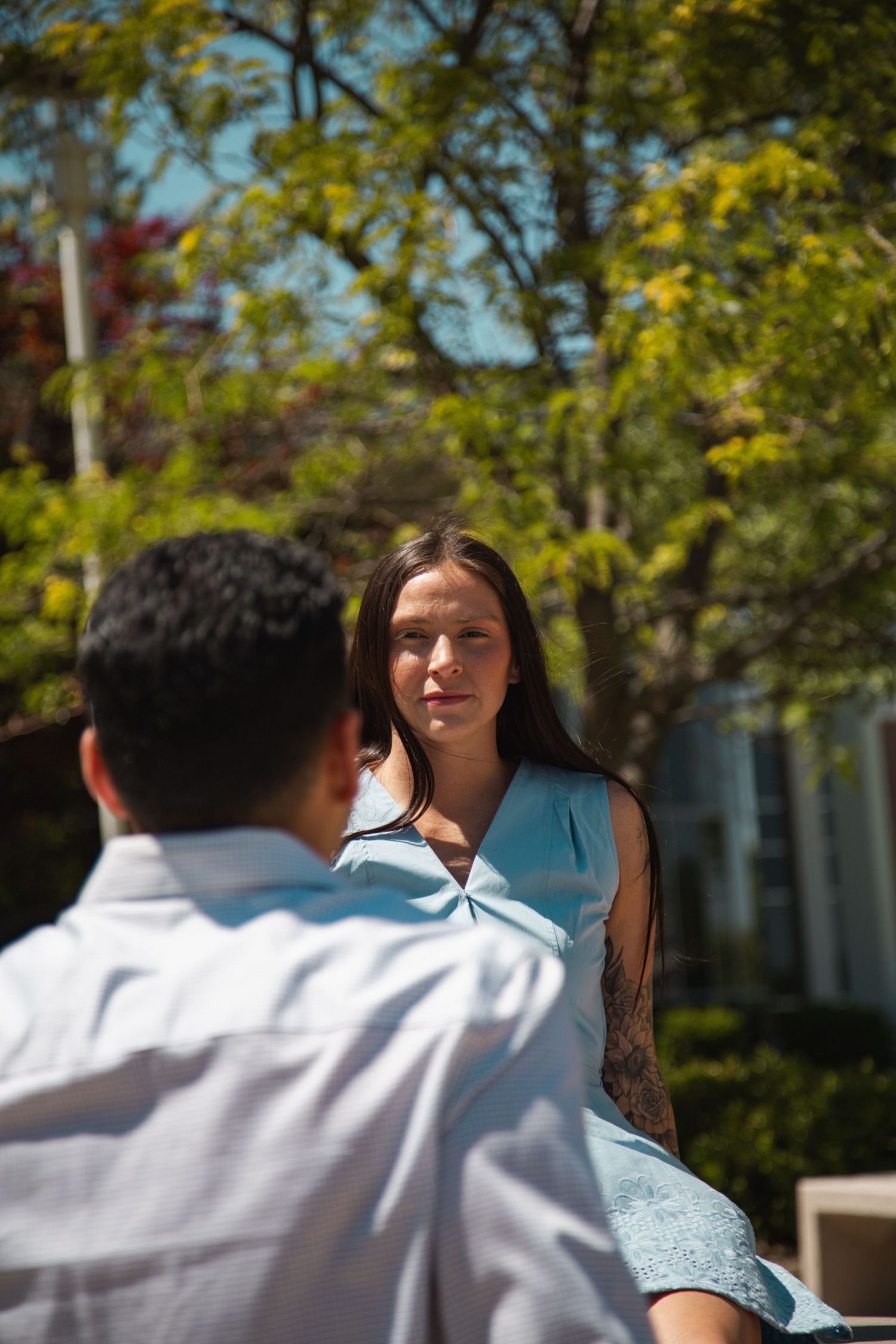 Young woman in blue dress sitting outdoors smiling at man in white shirt.