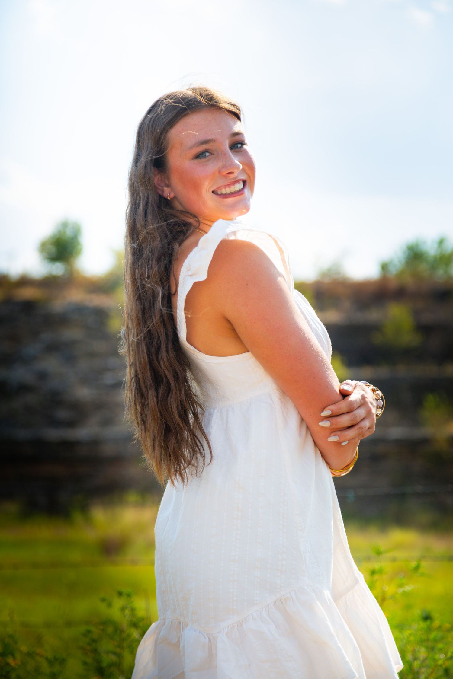 Young woman in white sundress smiling outdoors on green grass with stone wall background on sunny day.