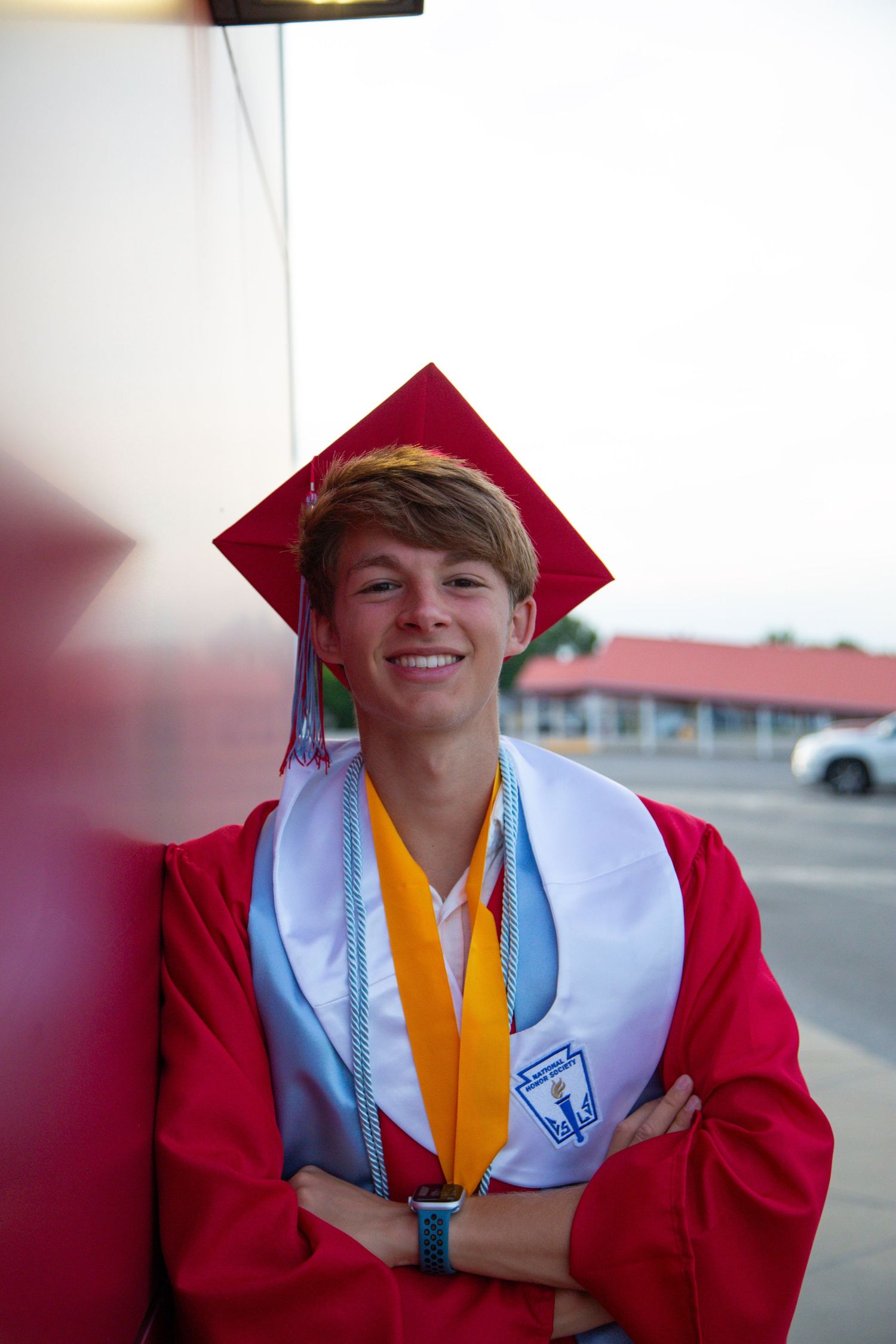 Smiling high school graduate in red cap and gown with gold and blue stoles poses with arms crossed.