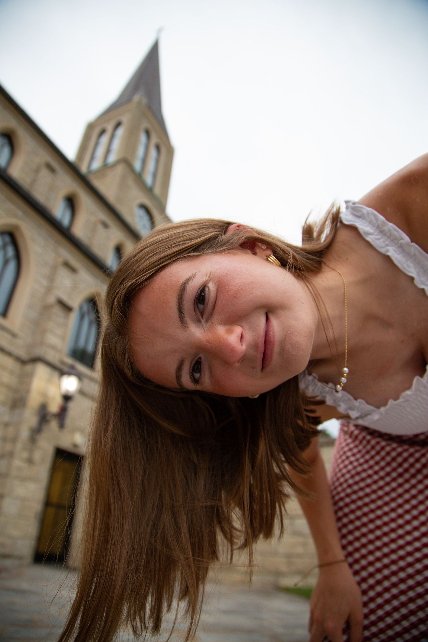Smiling teenage girl tilts head beside historic stone church with tall steeple.