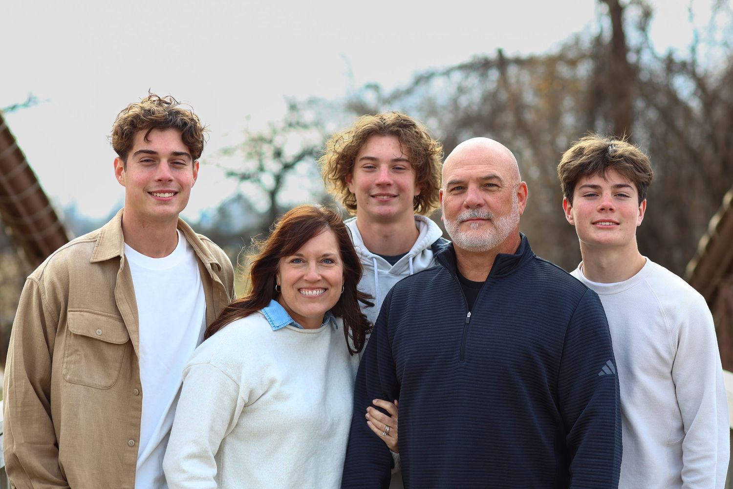 A happy family of five poses outdoors, with two parents and three teenage sons smiling together.