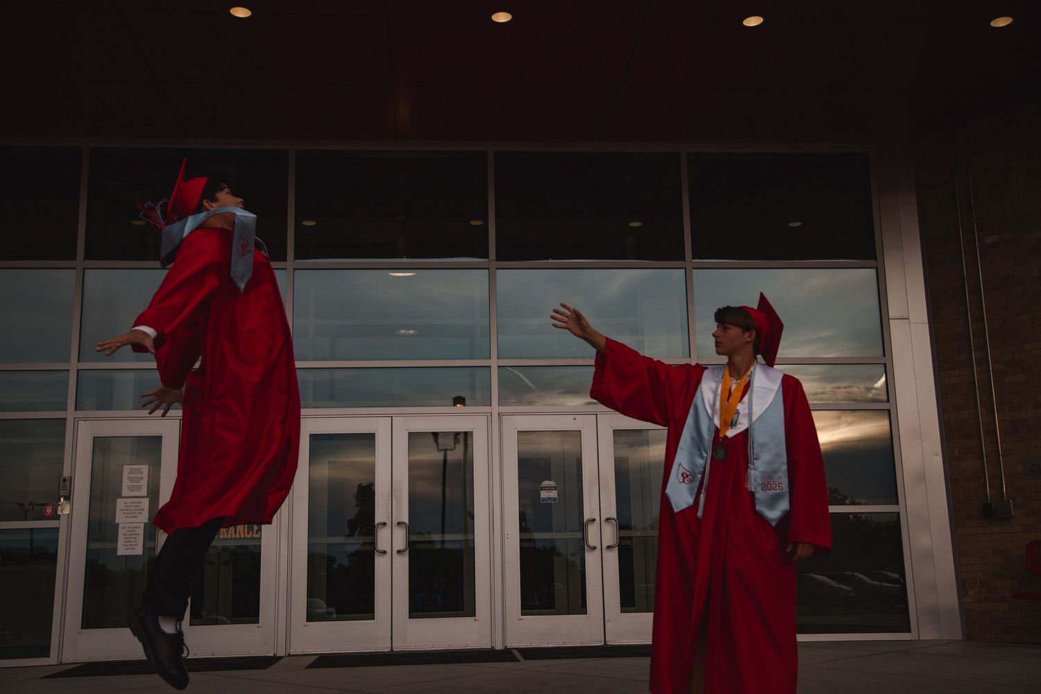 Two high school graduates in red caps and gowns, one jumping, outside a building at dusk.