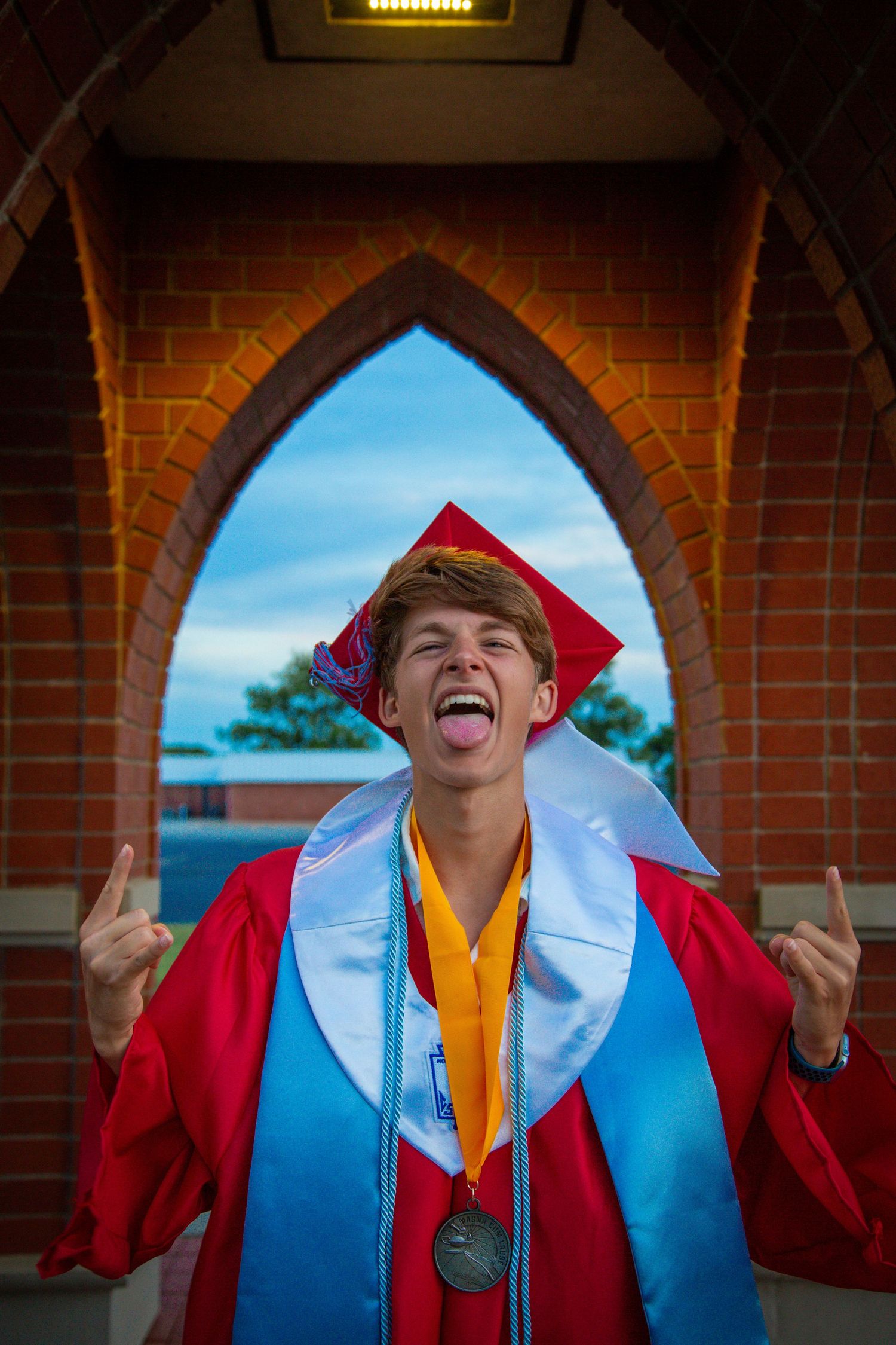 Excited high school graduate in red cap and gown making rock on gesture with tongue out under brick arch.
