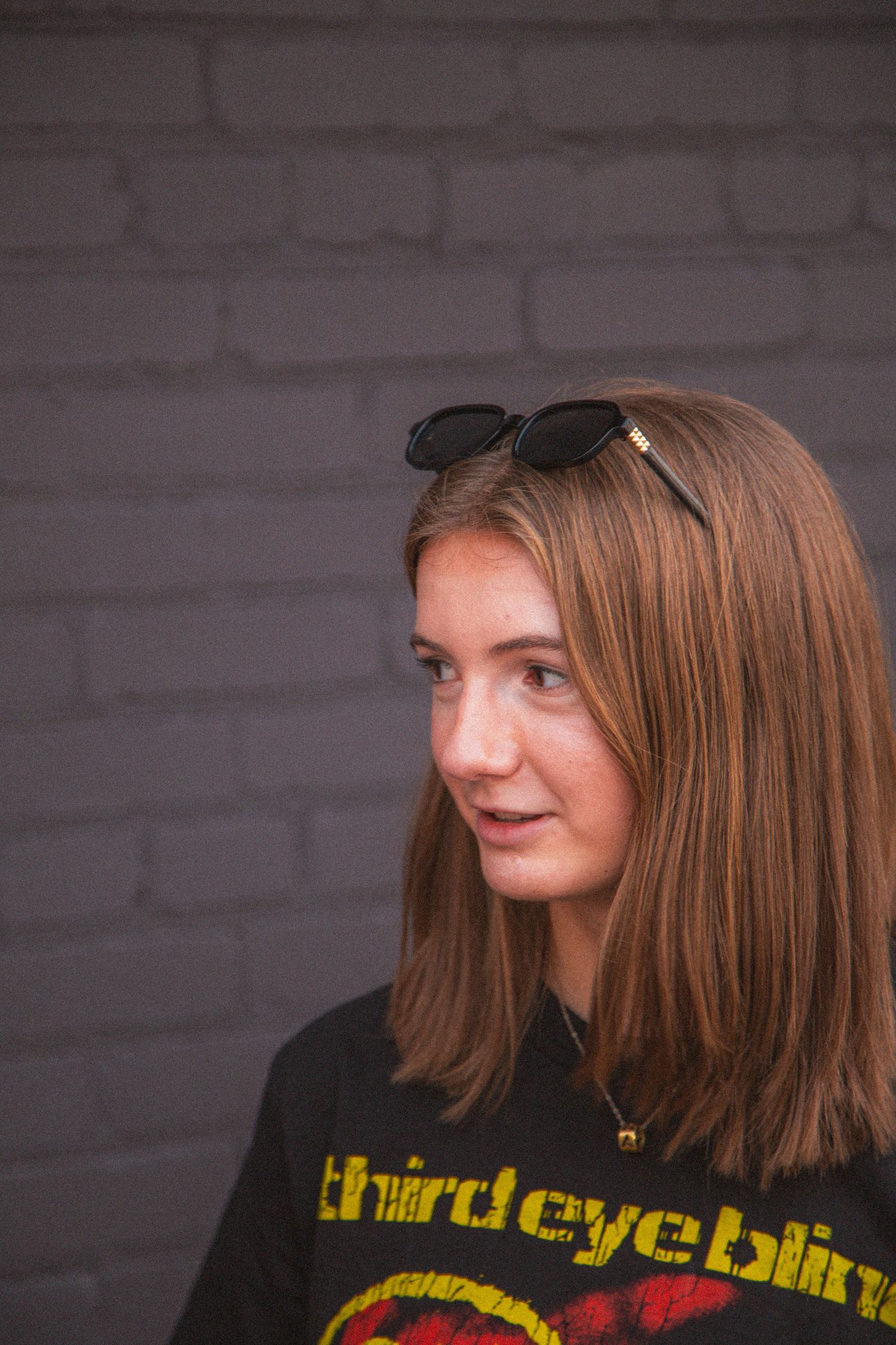 Young woman with sunglasses on head wearing Third Eye Blind shirt against gray brick wall.